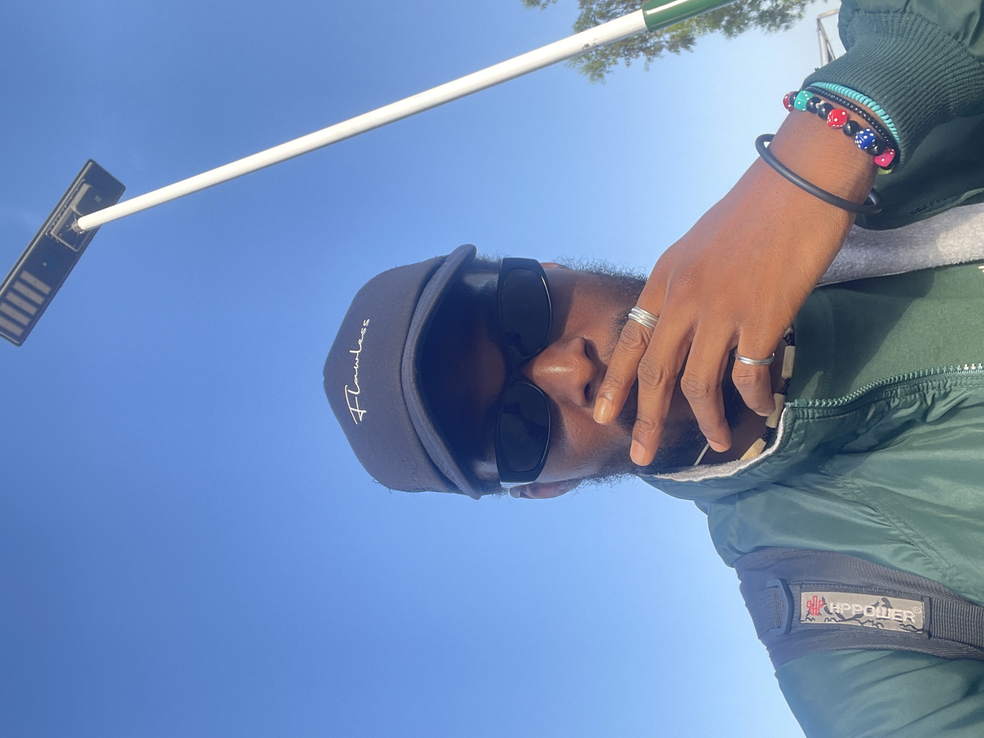 A low-angle shot depicts a person outdoors in Bar Jirgi Summa, Nigeria. The person, wearing a black baseball cap, sunglasses, and a green jacket with an "HPPOWER" patch on the sleeve, is looking down at the camera. They have bracelets on their wrist and rings on their fingers. Above the person is a bright blue sky with a solar-powered street light extending into the frame. Trees are visible in the far upper right corner.