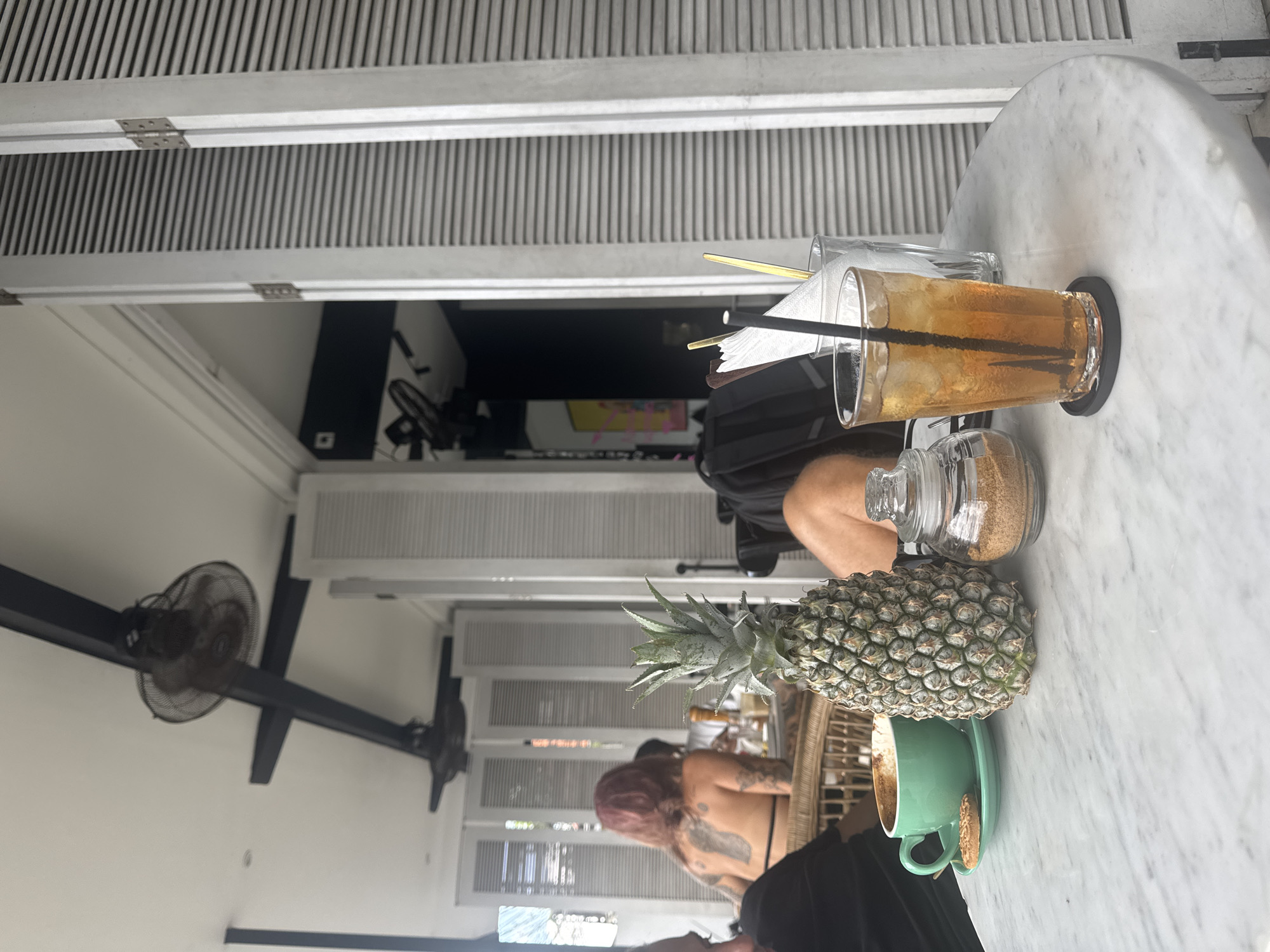 A high-angle, slightly rotated outdoor view shows a round marble table in what appears to be a cafe or restaurant setting. On the table, a glass of iced tea with a black straw and a black coaster sits adjacent to a glass jar filled with brown sugar. Further down the table is a whole pineapple and a partially consumed cup of coffee in a mint green cup and saucer, with a cookie on the saucer. Two people are present, though not in full view: one person with red hair and back tattoos is seated at a table behind the marble one, and another is seated at the marble table with their legs showing. The cafe has a casual ambiance, with white slatted walls visible in the background, along with a large fan on the left side of the view. The presence of the pineapple and relaxed atmosphere suggest a tropical or island-like setting. Based on the prompt, this is likely in North Kuta, Indonesia.