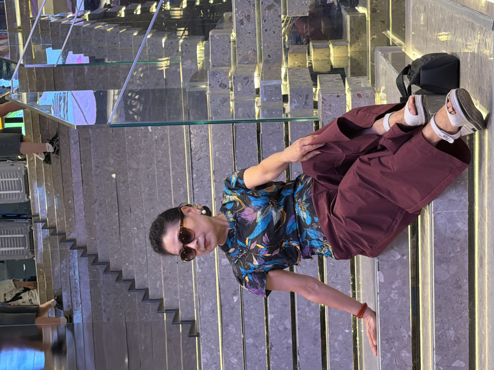 An Asian woman is seated on a wide, modern staircase in Yuzhong District, China. She is wearing a short-sleeved, patterned blouse in black, blue, and purple, and maroon wide-leg pants. White wedge sandals are on her feet, and a red beaded bracelet adorns her left wrist. She has dark hair pulled back, wears large brown sunglasses, and large pearl earrings. She is looking to her right, away from the camera. A black bag rests on the step behind her right leg. The staircase has broad, gray stone steps with a subtle speckled pattern and is illuminated by embedded yellow lights. A glass railing with metal supports runs along the right side of the stairs. In the background, other people are visible on the stairs, and the environment appears to be an indoor public space, possibly a mall or an exhibition hall, with reflective surfaces and bright lighting further up the stairs.