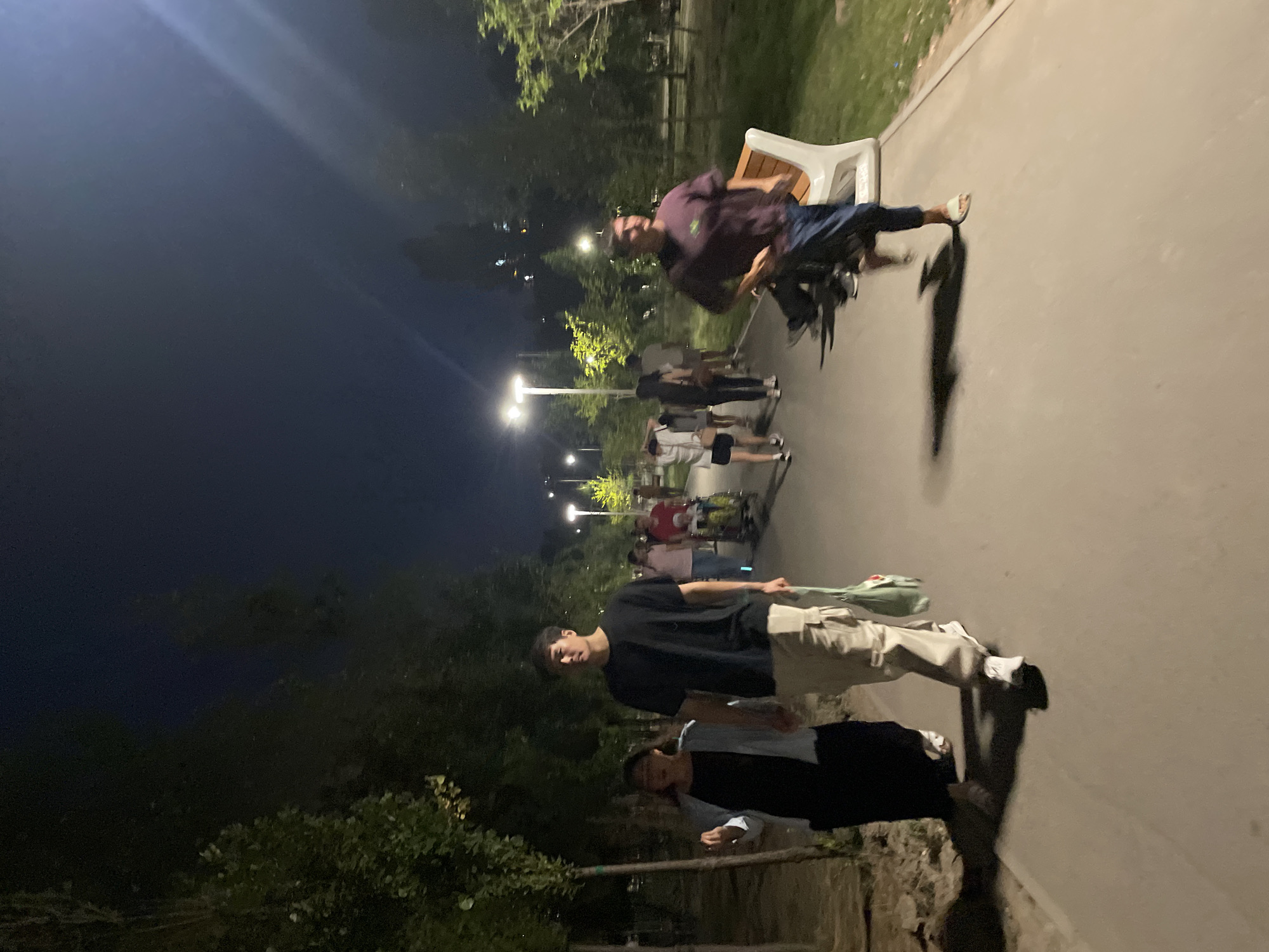 The image depicts a nighttime scene in a park or public walkway, likely in Almaty, Kazakhstan, given the provided context. Several individuals are present, walking along a paved path lined with trees. The lighting appears to be artificial, from street lamps, casting shadows on the ground.
In the foreground, a woman in a black dress and a light blue jacket walks hand-in-hand with a tall man in a black t-shirt and beige cargo pants. The man is carrying a light green bag. Behind them, further down the path, a group of people are visible, some walking, and one person appears to be pushing a stroller or cart. To the right, a man in a dark maroon t-shirt and blue jeans is walking past a modern-looking white bench. The background shows more trees and some distant city lights. The overall atmosphere is one of casual evening activity in an urban green space.