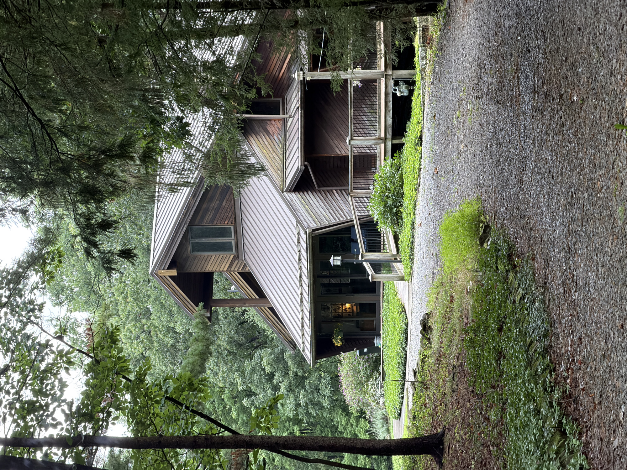 A rustic, multi-level wooden house is nestled within a dense forest. The house features a distinctive angled roofline and a wraparound wooden deck. A gravel driveway leads up to the house, which is surrounded by lush green foliage and mature trees. The sky is overcast, suggesting a cloudy or rainy day. No people are visible in the image. The location is identified as Wexford, United States of America.