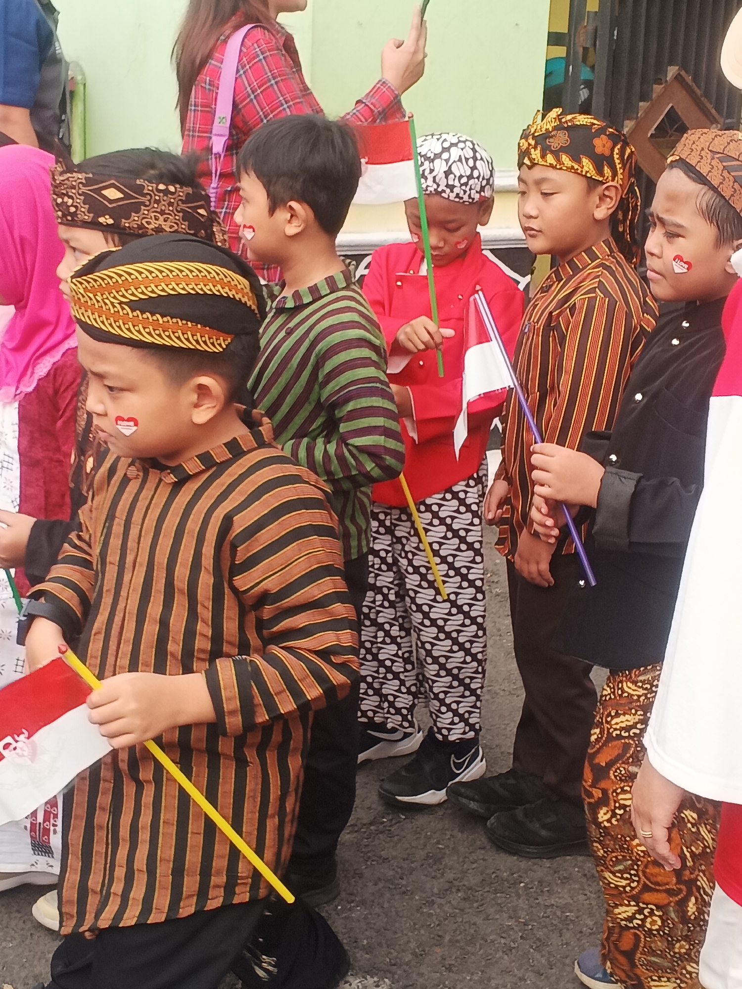 The image depicts a group of children, likely participating in a patriotic event, in Sokanegara, Indonesia. The children are dressed in traditional Indonesian attire, including batik patterns and head coverings. Many of them are holding small Indonesian flags, red and white, signifying the national colors. Some of the children have red and white face paint on their cheeks, resembling the Indonesian flag. The background shows a fence and a wall, suggesting an outdoor setting, possibly a public space or event venue. The overall atmosphere appears festive and celebratory.