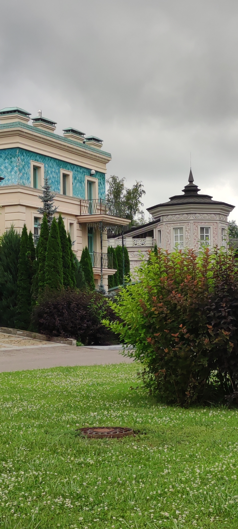 The image captures a view of two modern buildings within the Ангелово Клубная Резиденция in Russia, under an overcast sky. The building on the left is a multi-story structure with a light-colored facade adorned with turquoise patterned tiles on its upper section. It features several windows and balconies with decorative railings. A series of dark green, conical evergreen trees are planted in front of this building. To the right of this structure is a smaller, octagonal building with a domed roof, featuring decorative plasterwork and windows. In the foreground, a lush green lawn dotted with small white flowers leads towards a circular metal manhole cover. A variety of shrubs, some with green foliage and others with dark red leaves, are visible in the mid-ground and right side of the frame, partially obscuring the buildings and landscape. The overall scene depicts a well-maintained, upscale residential area with distinct architectural styles.