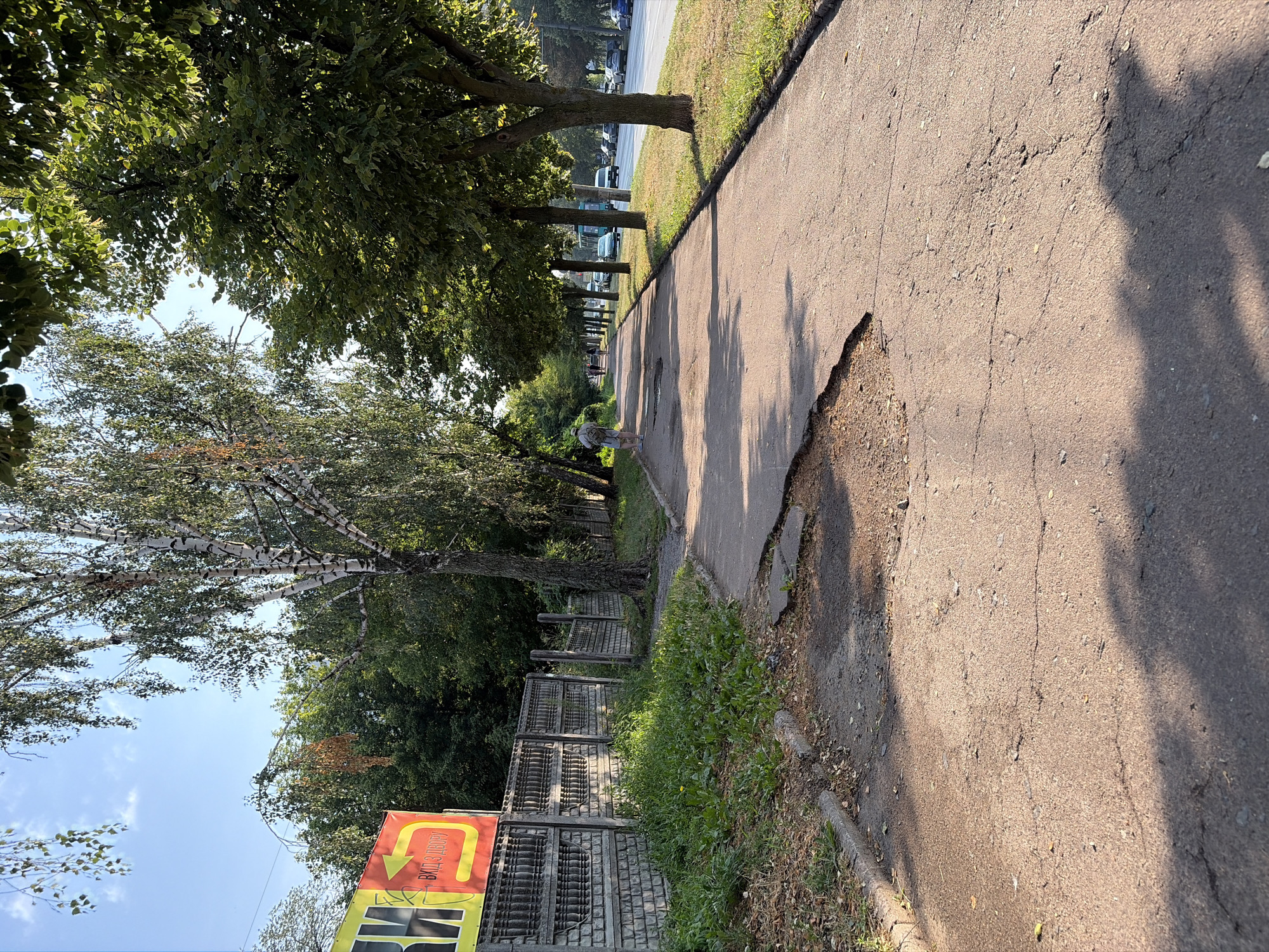 The image depicts a sun-drenched sidewalk in an urban environment. The sidewalk itself is in a state of disrepair, with a large section of pavement broken and uneven near the foreground. Cracks are visible throughout the surface. To the left of the sidewalk, there is a low concrete fence with decorative elements, bordered by a strip of overgrown grass and weeds. A yellow and red sign with Cyrillic lettering is partially visible behind the fence. Beyond the fence, dense green trees form a backdrop.
A person with a backpack and wearing a hat is walking away from the camera on the sidewalk, heading down the path lined with trees. Further down the sidewalk, several other figures can be seen walking, as well as a bus and cars on a road to the right. The trees lining the sidewalk cast shadows on the pavement, and sunlight filters through their leaves. The sky is clear blue with a few wispy clouds. The overall impression is of a typical summer day in a city, with a well-trafficked pedestrian path adjacent to a busy street.