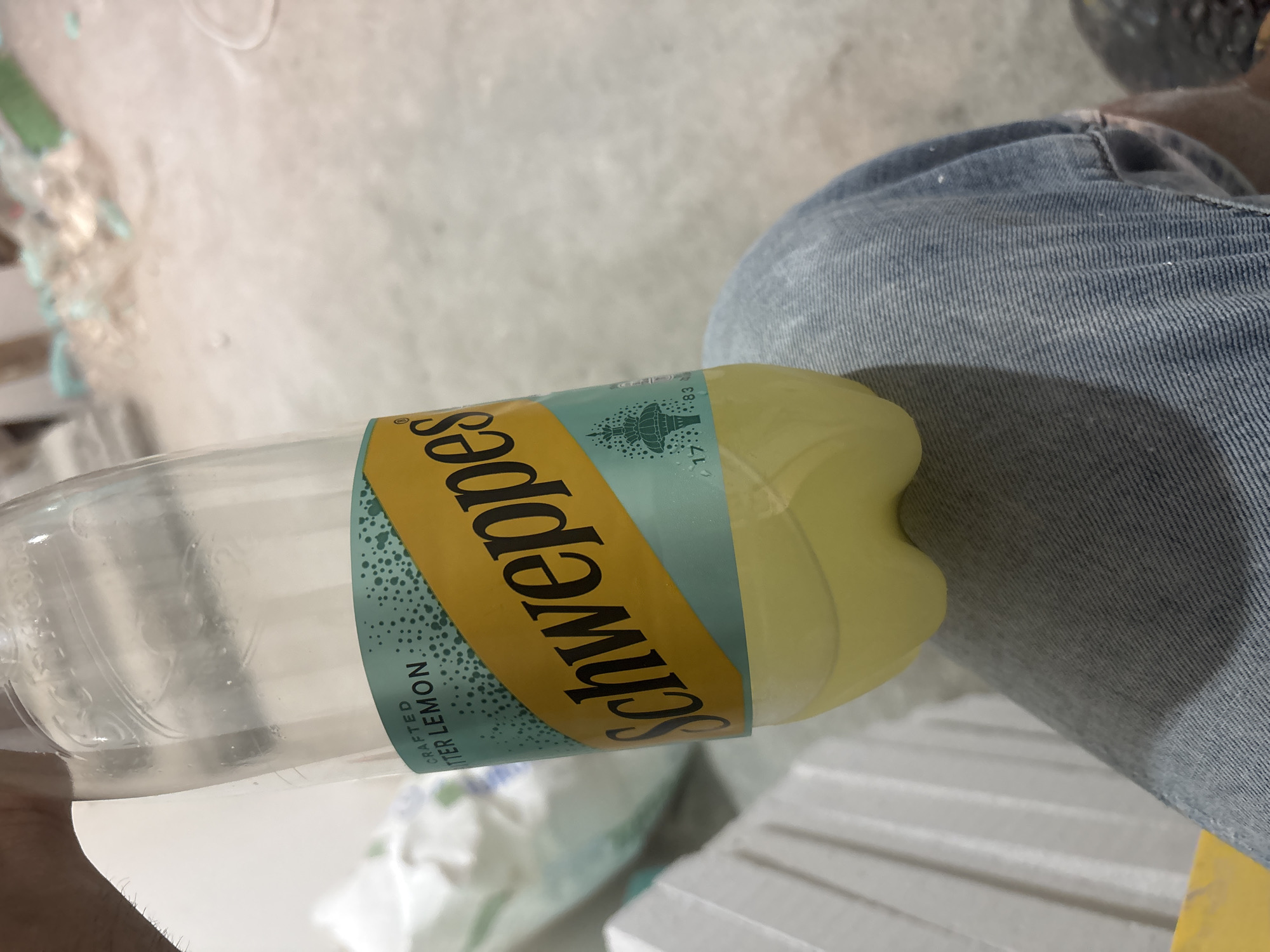A person is holding a bottle of Schweppes Bitter Lemon drink in a room under construction. The bottle is partially filled with a pale yellow liquid. The label on the bottle features the Schweppes logo and the words "CRAFTED BITTER LEMON". The background of the image is blurred, showing construction materials such as white panels and piles of debris. The floor appears to be concrete. The scene is indoors. There is no information in the image that would allow for the determination of a specific location within the city of Plovdiv, Bulgaria.