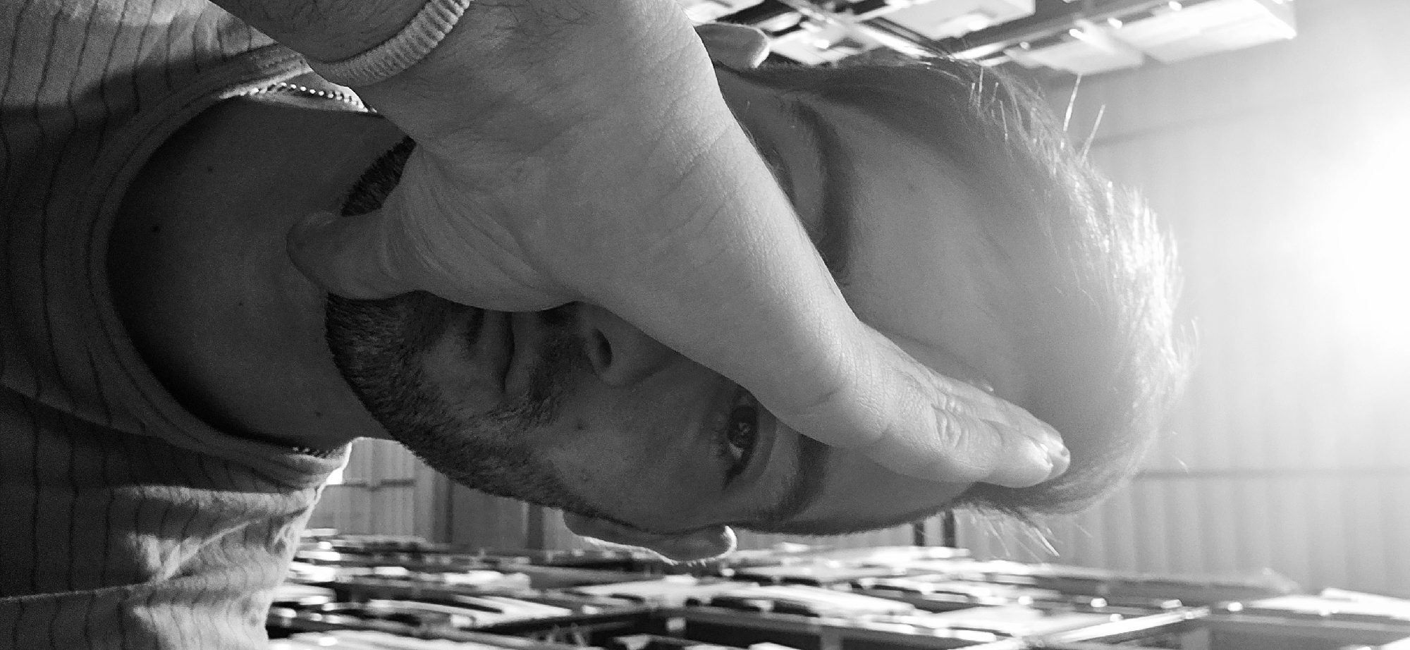 A black and white, close-up, upside-down shot shows a person with a hand covering the top half of their face. The person has a beard and a visible eye. The hand is wearing a bracelet. The person is wearing a striped shirt. The background is somewhat blurry but appears to show wooden planks. The location cannot be identified with the details available to be in the city of Plovdiv, Bulgaria.