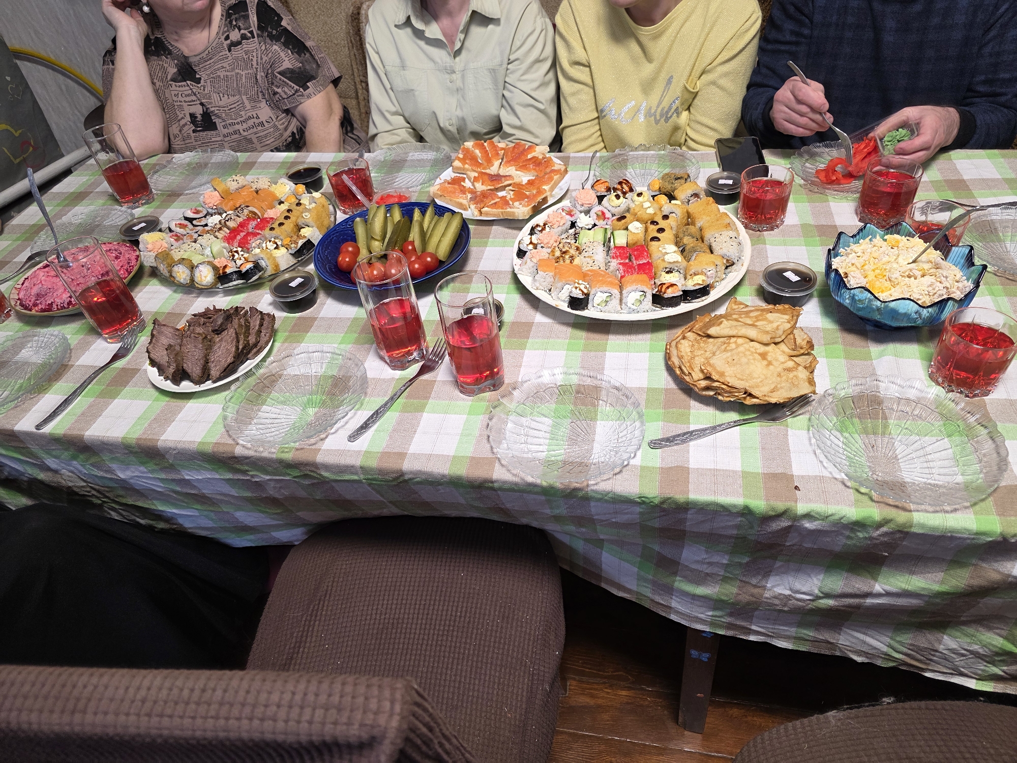 A group of at least four people is gathered around a table laden with a diverse meal, likely a family or social gathering in Chaikovsky, Russia. The table is covered with a green and white checkered tablecloth.
The subjects include a woman wearing a brown shirt with a newspaper print pattern, a person in a light green long-sleeved shirt, another in a yellow sweater, and a man in a dark blue sweater. The woman in the newspaper print top is resting her head on her hand, looking towards the table. The man in the blue sweater is actively using a fork to serve pickled ginger from a small container.
The table is densely packed with various dishes and drinks. Prominently featured are large platters of sushi and various types of rolls, including maki, nigiri, and tempura rolls. Other dishes include a plate of thinly sliced roast meat, a stack of blini (pancakes), and a platter of bread slices topped with red fish, likely salmon. There are also several salads: a blue bowl holds a creamy yellow salad, possibly Olivier or a similar chicken/cheese salad, and a clear bowl contains a pink/red salad, likely a beet-based dish. A blue bowl is filled with pickled cucumbers and cherry tomatoes. Small black containers of soy sauce and clear plastic containers of pickled ginger (both red and pale green) are interspersed among the dishes. Several clear glasses filled with a red beverage, possibly fruit compote or juice, are placed around the table.
The setting appears to be an indoor home environment, with what looks like a sofa and possibly some exercise equipment visible in the background. The general ambiance suggests a relaxed mealtime. The lighting indicates an indoor setting, likely evening or late afternoon.
Visible text includes partial newspaper headlines and articles on the woman's shirt, such as "of Cool," "el in October," "US Rejects Envoy," and "Provisions in." On the yellow sweater, a Cyrillic word, possibly "красивая" (krasivaya - beautiful), is visible.