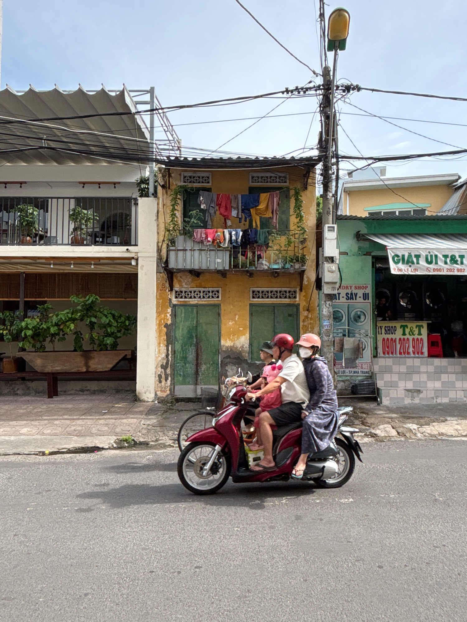 In Nha Trang, Vietnam, a red scooter carries three people along a paved street. The driver, wearing a red helmet and a white shirt, has a child in front of him wearing a pink shirt and a dark helmet. Behind the driver, a woman wearing a blue patterned robe and a pink helmet sits sideways, holding onto the driver. The scooter is passing by a building with a yellow facade and green doors. Clothes are hanging on a balcony above the doors. To the right of the yellow building is a laundromat with a green awning and laundry machines visible inside. A street lamp with a yellow top stands tall on a utility pole to the left of the yellow building, amidst a tangle of electrical wires. The sky is overcast.