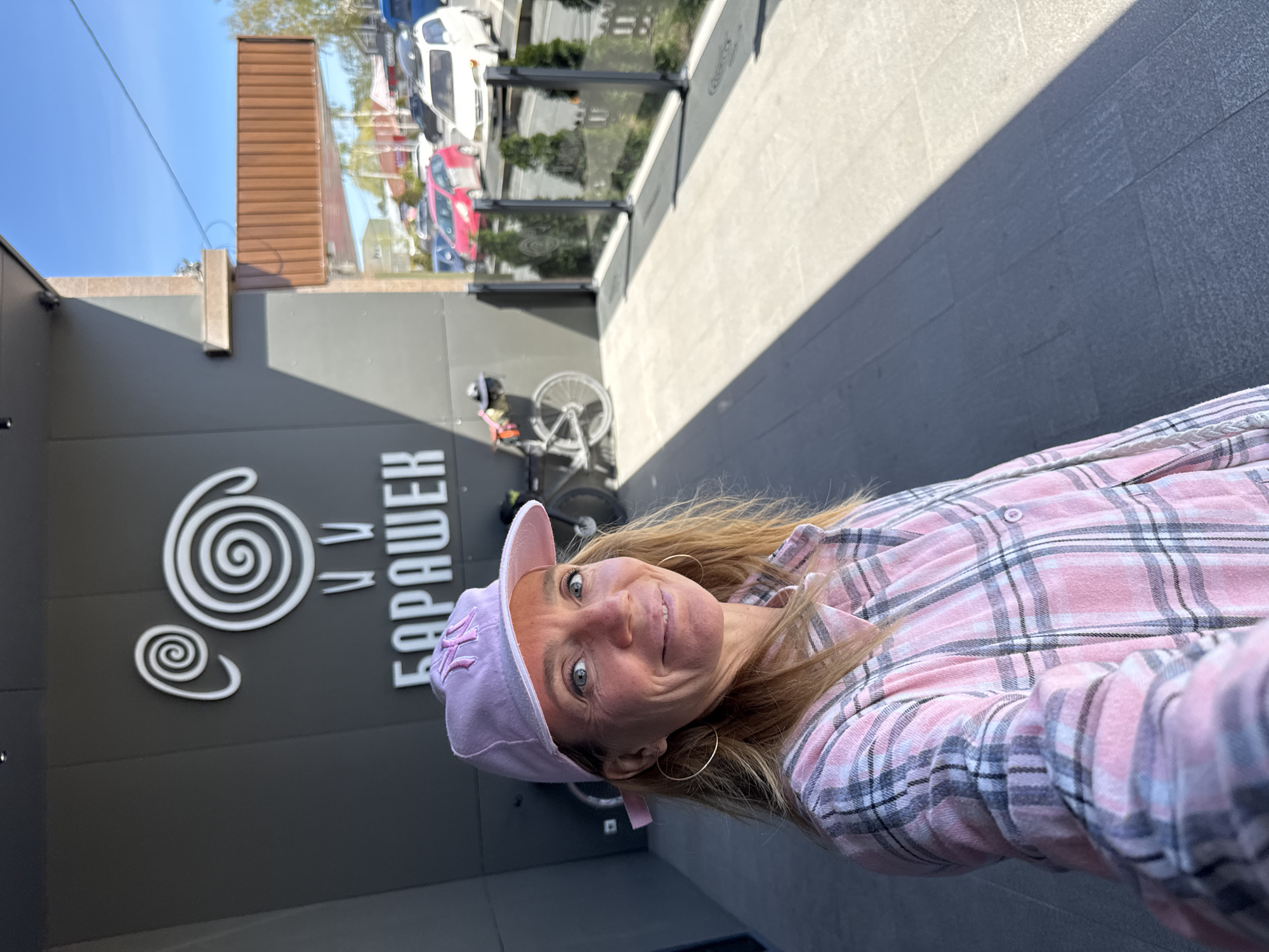 A woman in a pink plaid shirt and lavender baseball cap with the "NY" logo is taking a selfie outside a building in Çolpon-Ata, Kyrgyzstan. The building has a gray facade with a white sign showing a spiral design and Cyrillic letters spelling "Барашек". A bicycle is leaning against the wall near the entrance. The sky is blue, and sunlight casts a shadow on the pavement. Cars are visible in the background.