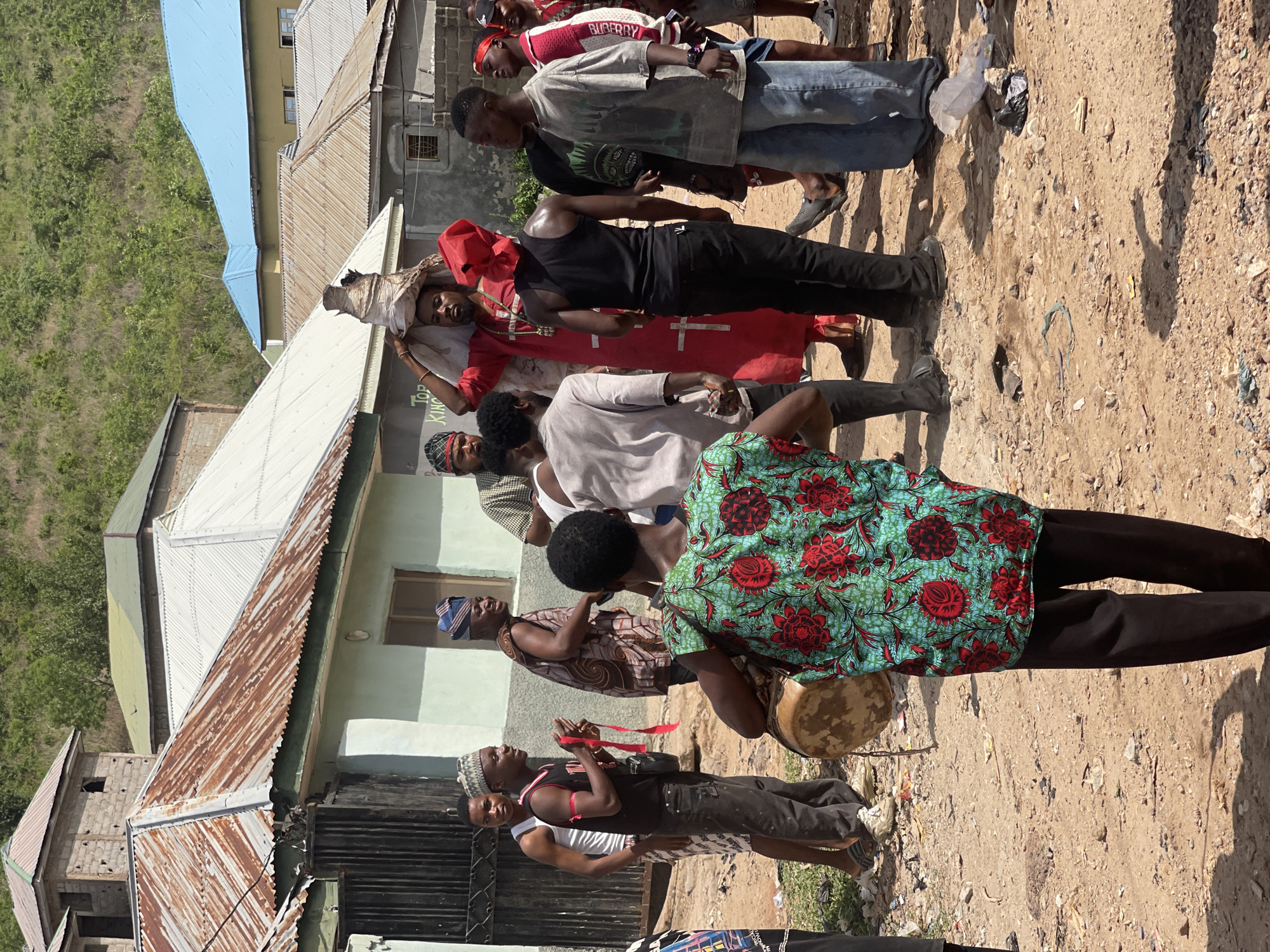 In Lokoja, Nigeria, a group of approximately ten individuals stand in a line along a dirt path next to a row of buildings with corrugated metal roofs. The people are diverse in age and attire. One person wears a green and red floral shirt and carries a drum. Another is clad in a red robe with a cross emblem, his head wrapped. Other subjects wear casual clothing. The surrounding environment includes buildings and vegetation.
