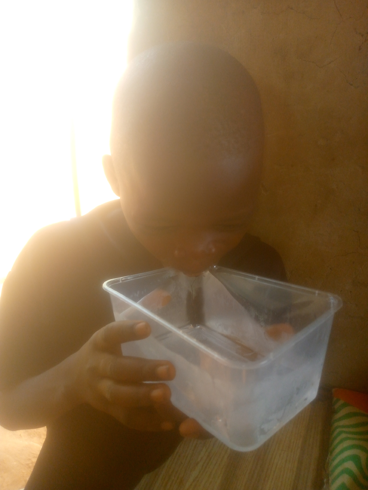A child is drinking water from a clear, rectangular plastic container. The child is facing forward, with their head tilted down to drink. The background is a blurred mix of light and shadow, with a textured wall visible on the right. The location and city are unknown.