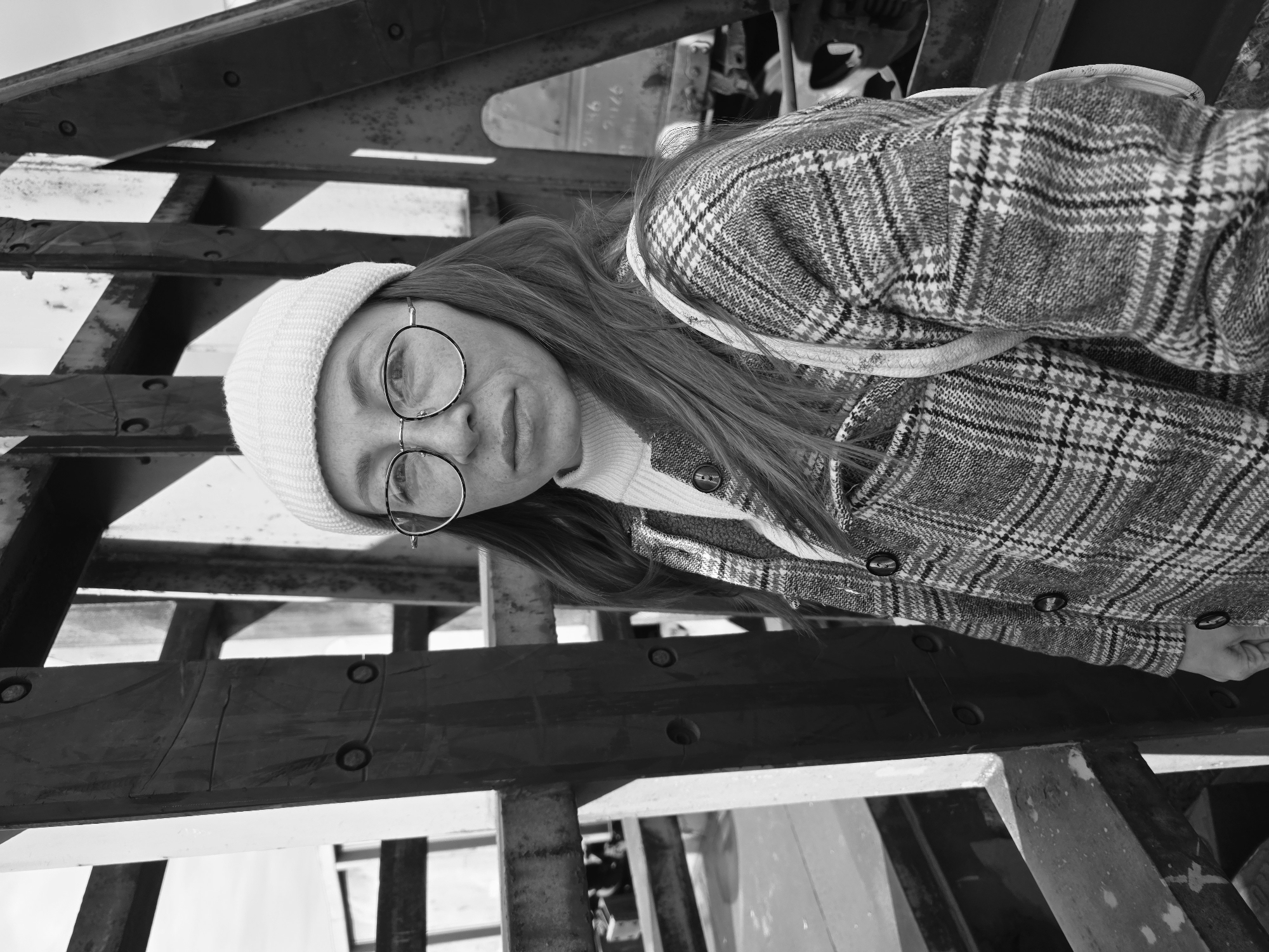 A black and white portrait features a young person, likely female, from the chest up, looking directly at the camera with a neutral expression. She wears a light-colored, ribbed turtleneck sweater, a matching knitted beanie, and a dark plaid jacket or coat with a houndstooth or similar pattern, secured by dark buttons. Distinctive round-rimmed glasses with thin metal frames rest on her face. Her long hair appears to be reddish-brown and falls over her shoulders.
The setting is outdoors, with the subject positioned in front of a substantial, angular structure composed of multiple beams, possibly metal or heavily treated wood, suggesting a bridge, pier, or industrial framework. Visible bolts or rivets are present on some of the beams, highlighting their construction. The background provides strong geometric lines and plays with light and shadow, creating depth. Faint, partially illegible numbers and text, possibly "91/16" and "2026," are visible on one of the background beams. The lighting suggests daylight, and the warm attire indicates cool or cold weather. The image has a direct and engaging quality. The location context is Chaikovsky, Russia.