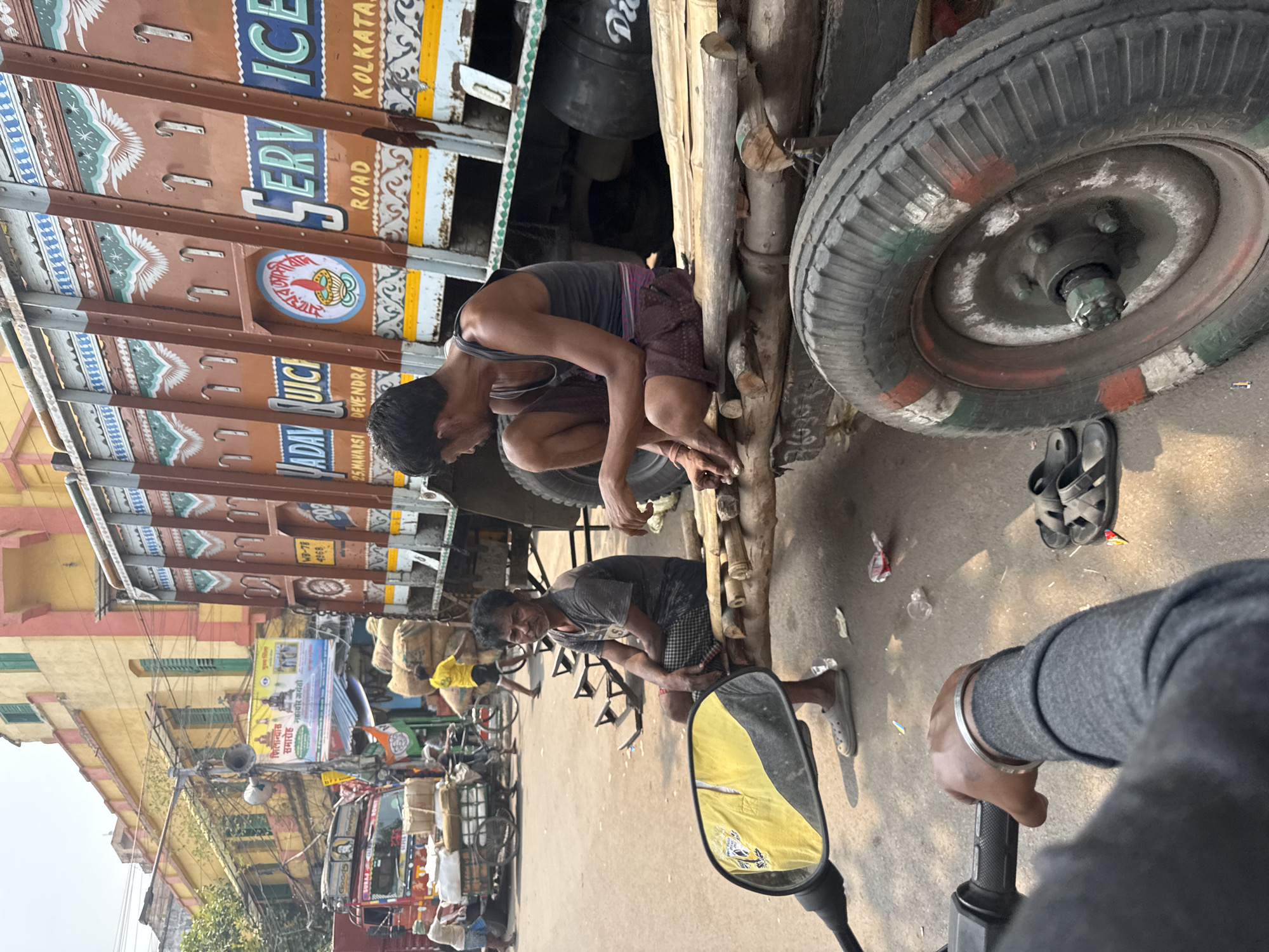 The image captures a street scene in Kolkata, India. Two men are squatting down, one near the front wheel of a large, ornately painted truck, and the other closer to the perspective of the photo, where we can see the side mirror of a motorcycle. The truck's side has lettering that reads "JADAV UICI, 25.MAHARSI DEVENDRA ROAD, SERVICE, KOLKATA." The truck is parked on a street with other vehicles and pedestrians. In the background, buildings can be seen with traditional Indian architecture and banners with Indian writing on them. The image is taken from the perspective of someone sitting on a motorcycle.