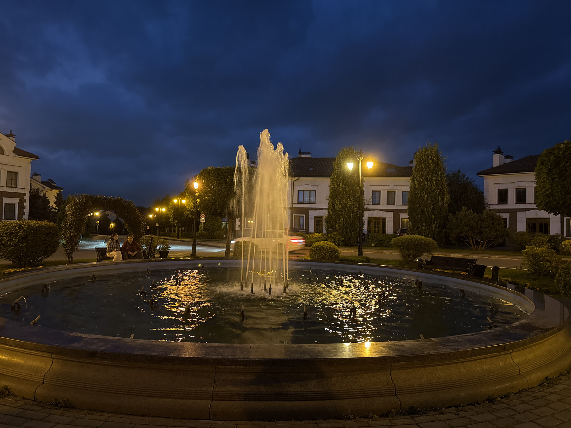 This is a wide-angle, low-light photograph taken at dusk in Lopatino, Russia. The scene is dominated by a circular fountain in the foreground, with water jets spraying upwards and illuminated by artificial light, casting reflections on the water's surface. The fountain is surrounded by a stone border. In the mid-ground, two individuals are seated on a bench beneath an arched green hedge. Behind them, a street is visible with lampposts and trees. Several multi-story residential buildings with traditional architecture line the street. The sky is a deep blue with visible clouds, suggesting the transition from day to night. The overall impression is a quiet, residential area at evening.