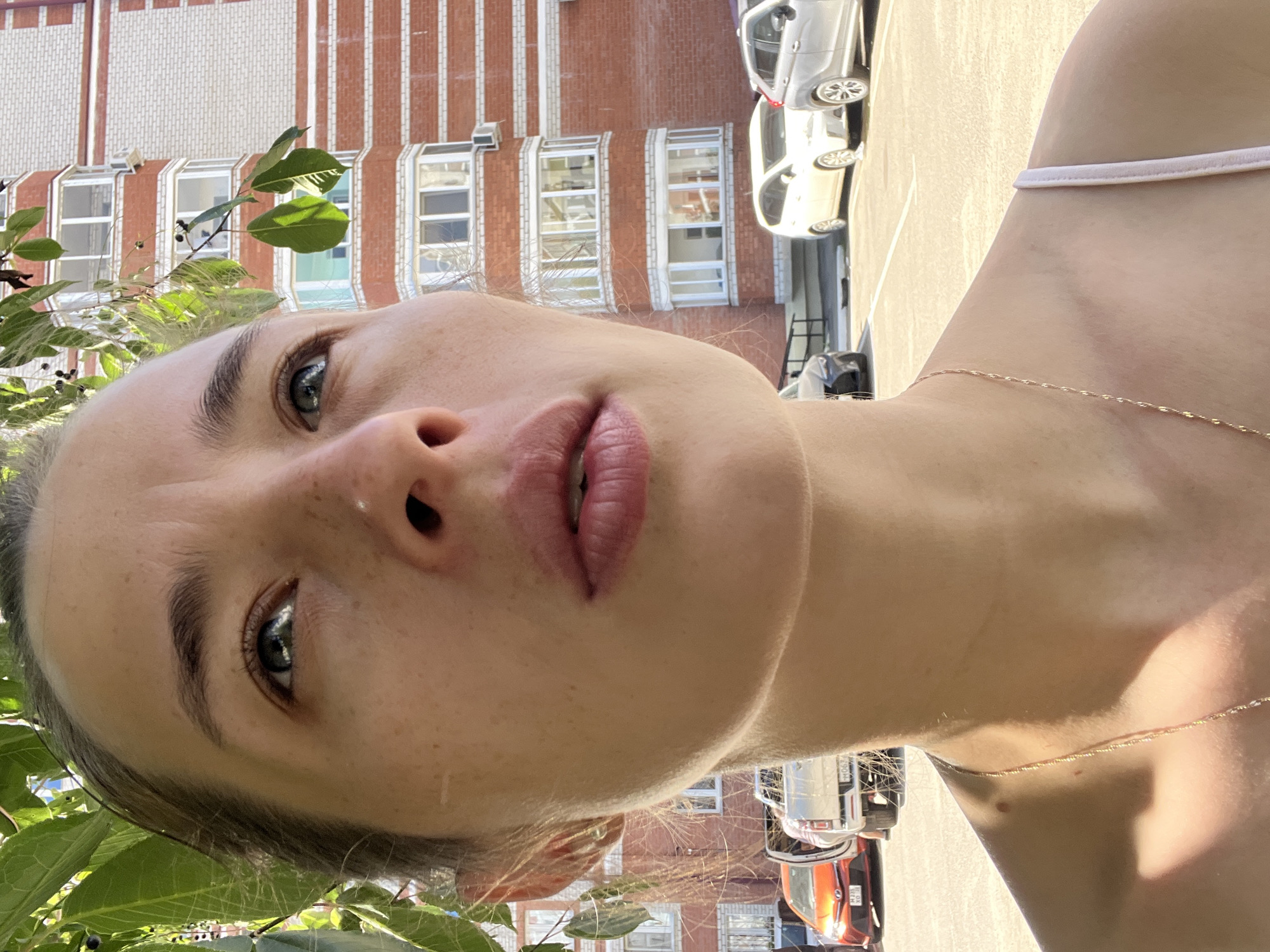 A close-up, chest-up, outdoor, medium shot captures the face of a young woman looking upwards, with a building and parked cars in the background. The woman's face is partially obscured by green leaves and branches from a tree. She has light skin with freckles and a small mole on her cheekbone. Her eyes are blue-gray, and her lips are slightly parted. She is wearing a thin, light-colored strap, possibly from a dress or tank top, and a delicate gold chain necklace. The background features a modern, multi-story building with red brick and white trim, and several cars parked in a lot. The lighting suggests it is a sunny day. The provided information that the image is located in Chigiri, Russia, is not visually verifiable from the image itself.