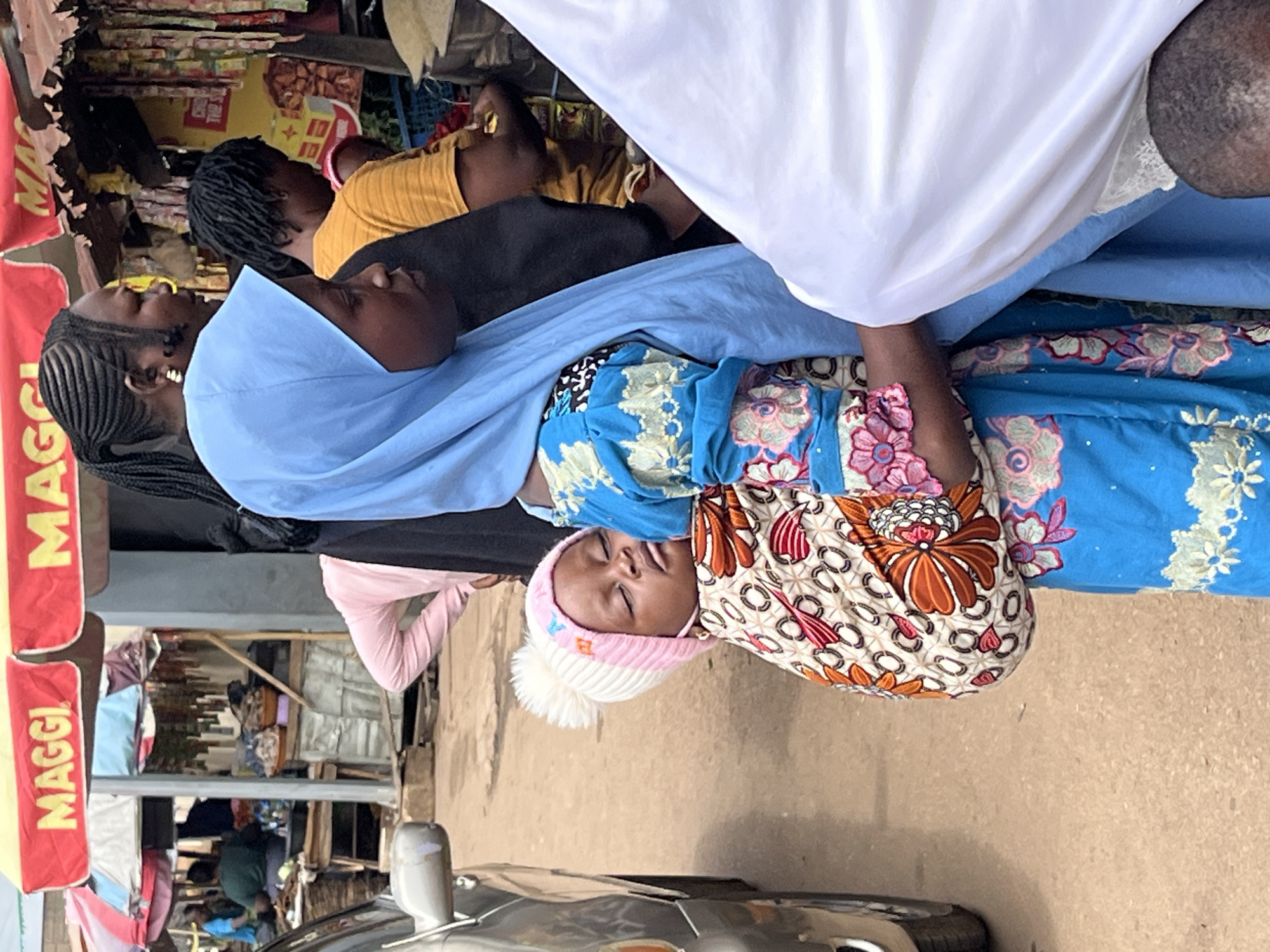 In Jos, Nigeria, a woman wearing a blue hijab and a colorful blue and floral patterned dress carries a baby in a patterned fabric sling. The baby, wearing a pink and white pom-pom hat, sleeps soundly against her mother. Behind them, other people are visible at a market stall under red awnings displaying "MAGGI". A silver car is parked to the left of the scene. The environment suggests an outdoor market setting.