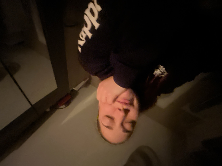 A person wearing a dark hooded sweatshirt with white writing is pictured indoors. The person's eyes are closed, and their hand is touching their face. A slender object with a red handle and silver blade is seen along the wall. The lighting is dim, and the photograph is oriented upside down. A partial view of a pale wall and floor are also present.