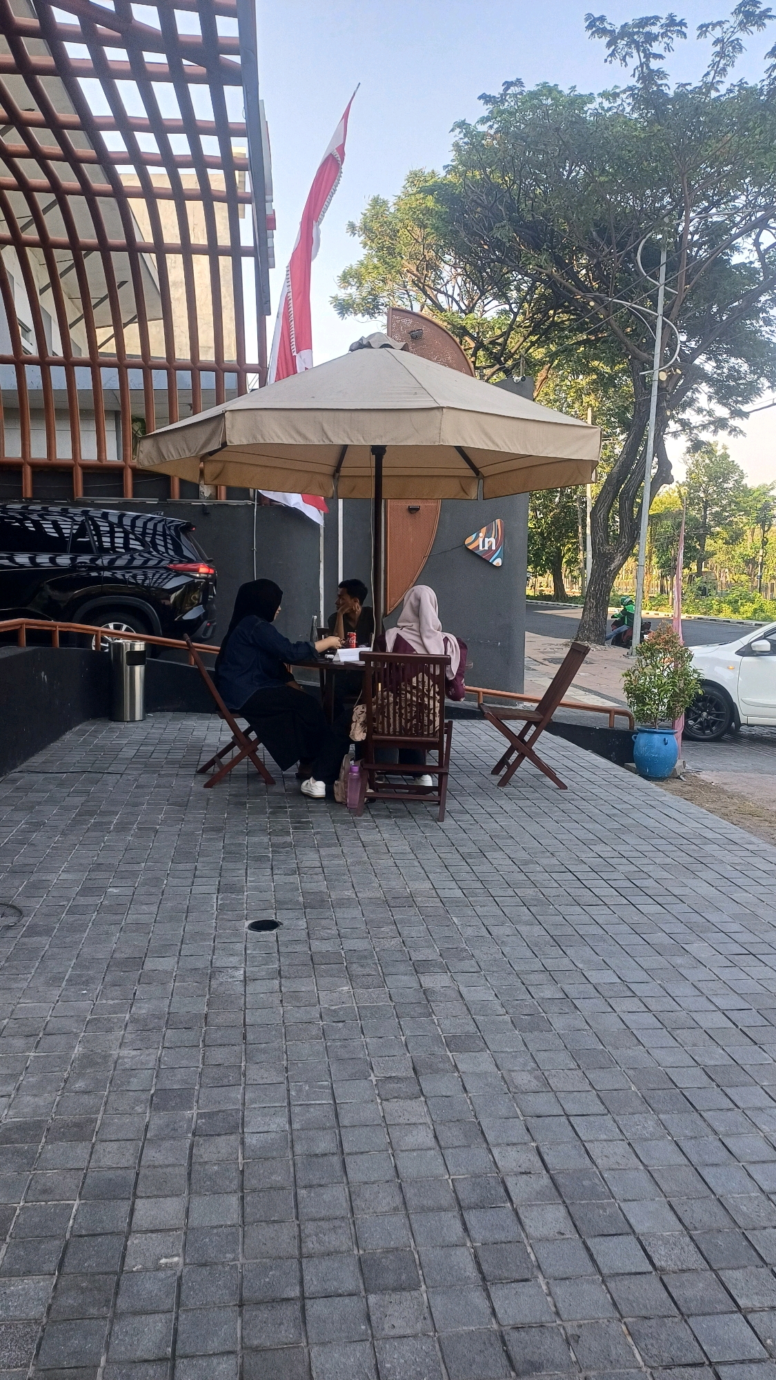 In Surabaya, Indonesia, on a bright, clear day, three individuals are seated at a table outdoors, under a large beige umbrella. The table is round and dark. Two women, both wearing hijabs, are seated on one side. The woman on the left is wearing a dark blue denim top and black pants, and appears to be gesturing with her left hand while looking at the person across from her. The woman on the right is wearing a maroon and beige patterned hijab and a maroon top, and is looking down at the table. A man with dark hair is seated between them, facing the woman in the blue top and wearing a dark green shirt. He is leaning forward, resting his chin on his hand. On the table, several items are visible, including what appears to be a drink and a phone.
The seating area is paved with grey square cobblestones. To the left of the table, a black SUV is parked. A tall, metallic trash can is positioned next to the SUV. Behind the group, the exterior wall of a building is visible, painted in dark gray with a curved, reddish-brown architectural element. A small sign with an "in" logo is affixed to the wall. In the background, a large tree with dense green foliage provides shade. A white car is parked to the right, and a blue pot containing a small plant sits on the cobblestone path. A street is visible beyond the plant and the white car, with a person on a motorcycle in the distance. A red and white flag is prominently displayed on a flagpole near the building.