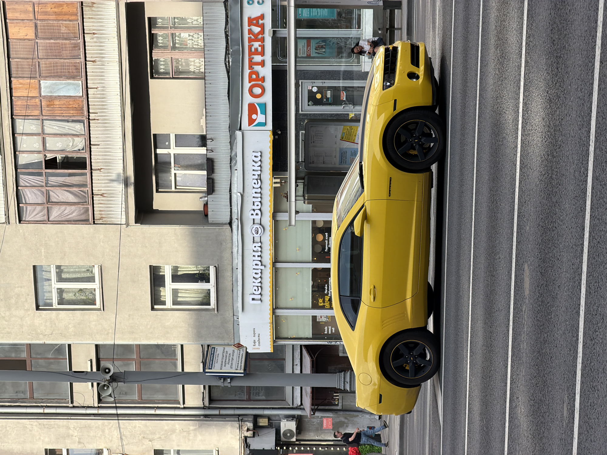 The image shows a yellow Chevrolet Camaro parked on a city street in Moscow, Russia. In the background, a building with multiple windows and balconies is visible, along with storefronts for a bakery and a pharmacy. A street lamp with three speakers is also present. A man walks on the sidewalk, and a woman sits on a bench near the pharmacy. The street has multiple lanes with white markings.