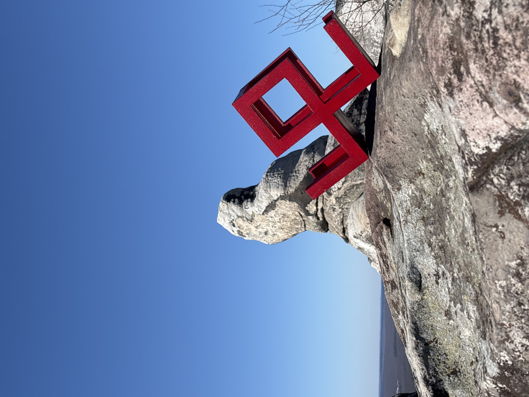 A red metal sculpture sits precariously on a rock formation against a clear blue sky. The sculpture is composed of interconnected square and rectangular frames, forming an abstract, geometric design. The rock formation appears to be part of a larger cliff or mountain face, composed of gray and brown stone. The image is taken at such an angle to make the sculpture and rock appear to be on the side. This photograph depicts the "Mountain Cross" landmark, an art object by Perm sculptor Aleksei Zavyalov, located in Gubakha, Russia.