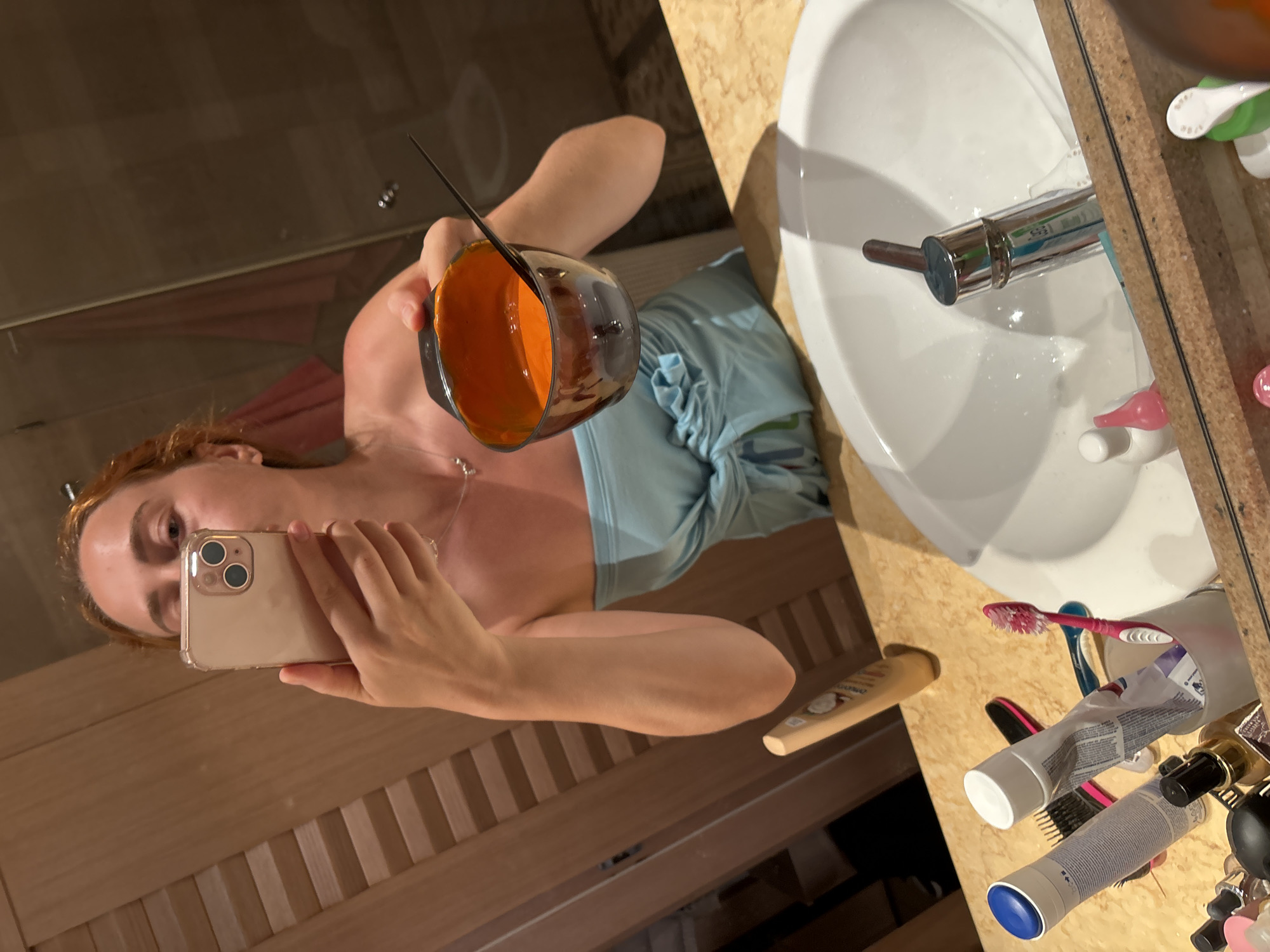 The image depicts a woman in a bathroom, taking a selfie in a mirror. She has red hair and is wearing a blue strapless top. She is holding a phone in her right hand and a small bowl containing an orange substance with a black applicator in her left. The bathroom has wooden paneling on the walls and a granite countertop with a white sink. Various toiletries, including toothpaste, toothbrushes, and bottles, are visible on the countertop. The scene suggests the woman is preparing to apply hair dye. The location is identified as Budva, Montenegro.