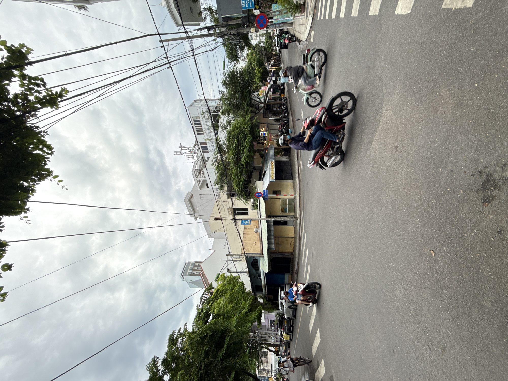 In Nha Trang, Vietnam, on a cloudy day, a street intersection is depicted. Several motorbikes are on the road, with riders wearing helmets. In the foreground, a person on a red motorbike is riding towards the right, and a person on a light blue motorbike is riding towards the left. In the middle ground, a person on a red motorbike is riding towards the viewer, and another person on a light green motorbike is riding towards the viewer. Beyond the intersection, buildings line the street, some with awnings and signs. Trees and overhead power lines are visible. A pedestrian crossing is painted on the asphalt road.