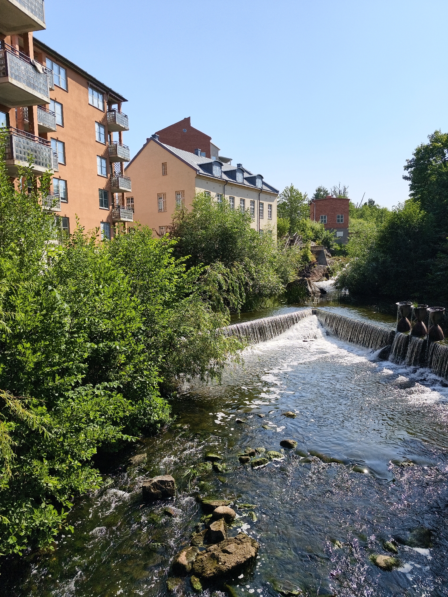 The image captures a scenic view of the river Stångån flowing through Linköping, Sweden. On the left, a modern residential building with multiple balconies is visible, constructed with orange-toned plaster and white balconies. Adjacent to it, a smaller, older building with a similar orange hue and arched windows stands. Further in the background, a red brick building is nestled amongst lush green trees. The river itself features a man-made waterfall, with water cascading over a series of steps and several cylindrical structures on the right bank. The water is clear, with visible rocks and vegetation along the riverbed. The scene is bathed in bright sunlight, indicating a clear day.