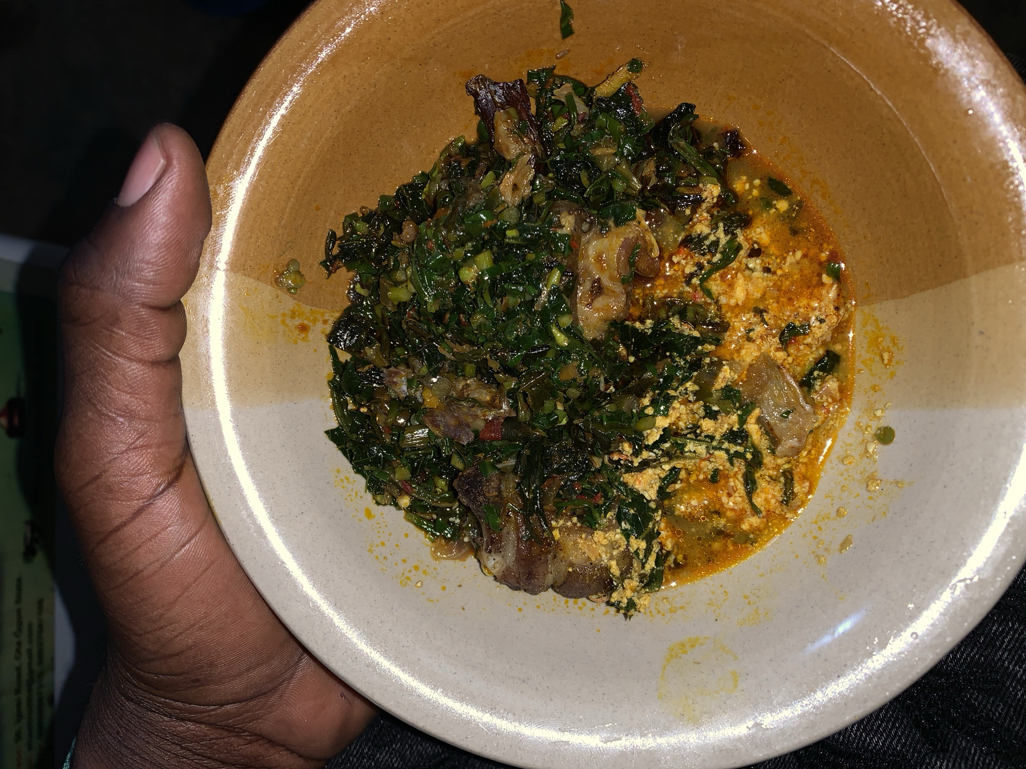 A dark-skinned person is holding a bowl filled with food. The bowl is ceramic, with a beige interior and a tan exterior rim. The food consists of two distinct sections: one is a leafy green vegetable dish, possibly a stew, with what appears to be pieces of meat or fish mixed in; the other is a yellow, granular mixture in a light orange sauce. The person's thumb is visible on the left side of the image. The setting appears to be indoors. There is no information provided in the image to corroborate the city of Kabong, Nigeria, or any indication of a specific action or interaction taking place.