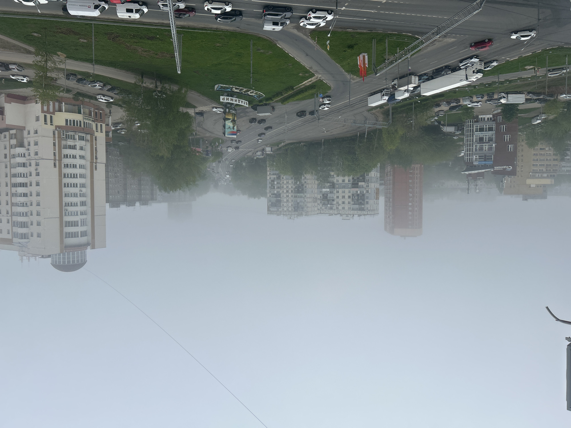 The image shows a city scene with buildings, roads, vehicles, and greenery, all viewed through a thick layer of fog. The buildings vary in height and architectural style. Roads intersect, with vehicles lined up on one side and moving along the other. Green areas are interspersed with the buildings and roads. The fog obscures details and creates a muted, gray overall tone. The location appears to be in Vladimir, Russia, based on a sign visible in the image.