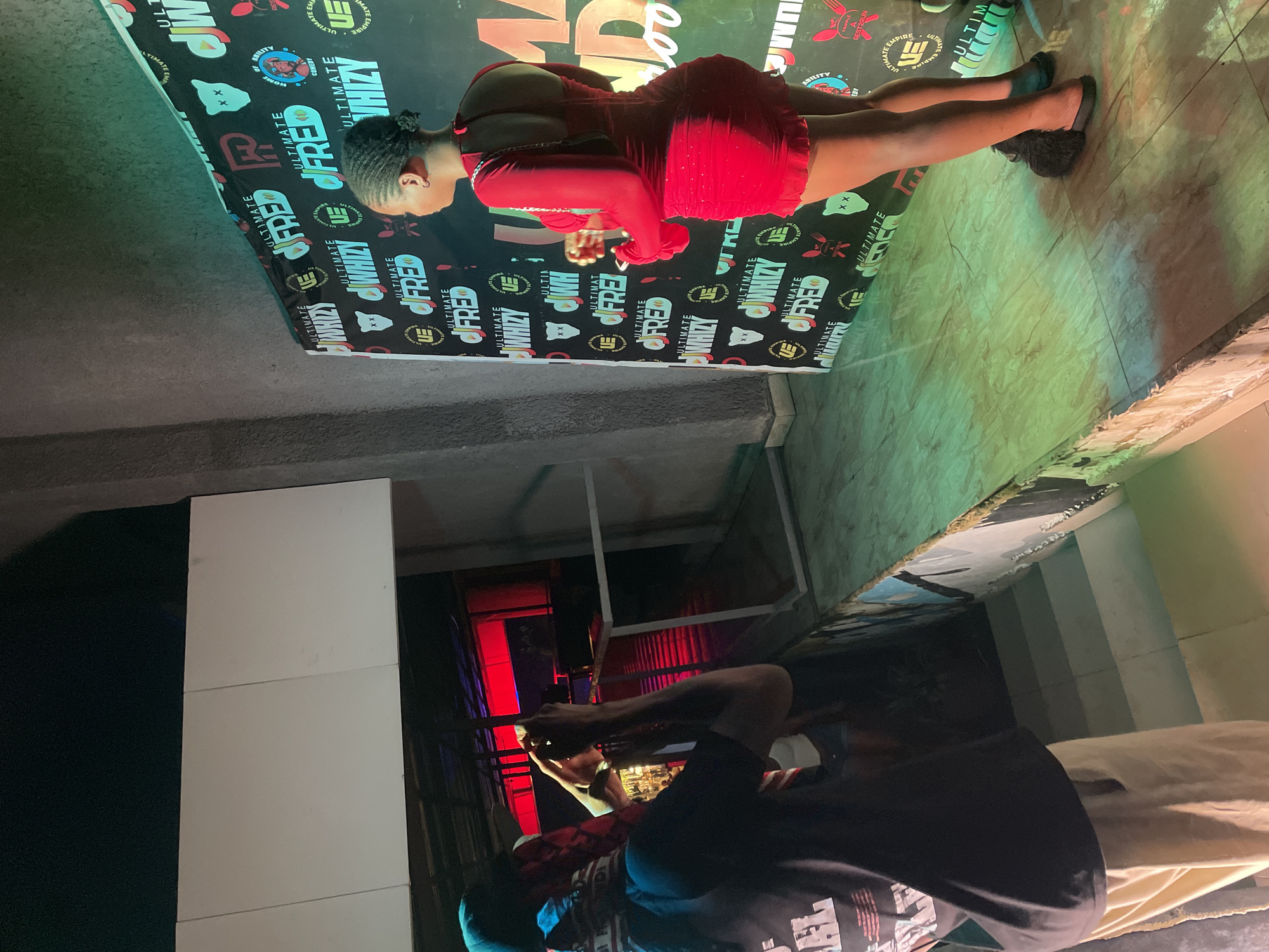In Kurunduma, Nigeria, a young woman in a red dress poses for a photograph against a backdrop featuring the logos of "Ultimate Whizy" and "Ultimate Fred." She is positioned on a stage illuminated by green and yellow lights, with a photographer on the left capturing the shot. The photographer, wearing a black t-shirt and a blue head covering, is holding a smartphone up to take the picture. The backdrop is dark and features repeating graphic elements, including stylized bears and the aforementioned brand names. Behind the photographer, a staircase leads down into a dimly lit area with red lighting. The overall setting appears to be an indoor venue, possibly a club or event space, with a concrete wall on the left and a textured floor.