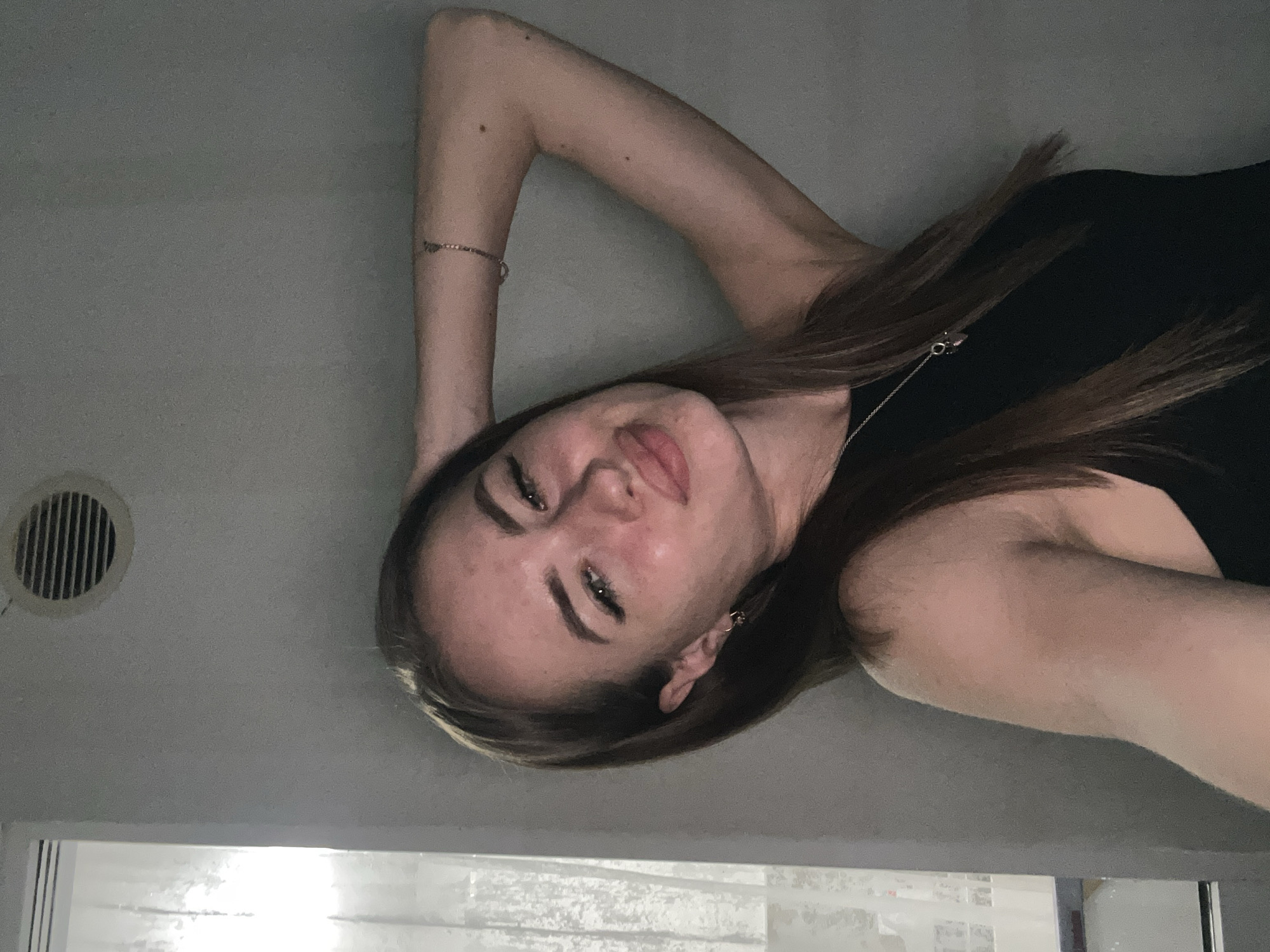 In an indoor setting, a young Caucasian female with long brown hair is captured in a selfie-style portrait. She is wearing a black sleeveless top and a silver necklace. Her right arm is bent and resting on her head, and she is looking towards the camera with a slight smile. A small tattoo of a heart with an arrow through it is visible on her right upper arm, just above a thin bracelet. The background consists of a plain gray wall with a circular vent in the upper left corner. To the left of the subject, a sheer white curtain is visible, suggesting a window. The lighting appears to be artificial. The location is not specified.