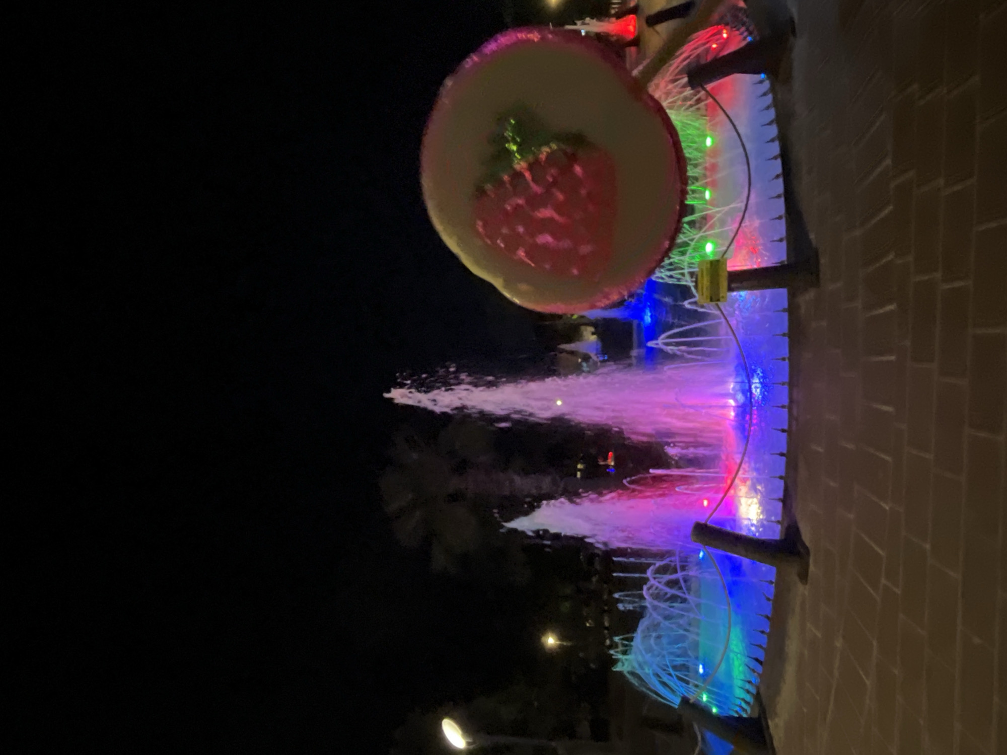 An outdoor night scene, possibly in Alanya, Turkey, features a vibrant and colorful illuminated display. The focal point is a large, decorative, round object resembling a stylized strawberry or fruit slice, glowing with a pink center, yellow rim, and green accents. This brightly lit decoration appears suspended or mounted above a multi-tiered water fountain.
The fountain itself is dynamically illuminated with an array of colors, including vivid blues, purples, pinks, and greens. Streams of water shoot upwards, catching the colorful lights and creating a lively visual effect. The fountain area also incorporates structural elements and possibly railings, adorned with small, colorful LED lights that contribute to the festive atmosphere.
To the right side of the fountain, a light brown brick wall is visible, suggesting a pathway, building facade, or landscaped area bordering the display. The background is predominantly dark, indicating nighttime, with faint glimmers of distant lights and indistinct shapes that could be trees or other urban structures. No individuals are visible in the image. A small, indistinct yellow object, possibly a sign or part of the lighting apparatus, is present near the green lights of the fountain structure, but no legible text can be discerned. The overall impression is one of a decorative public light and water show.