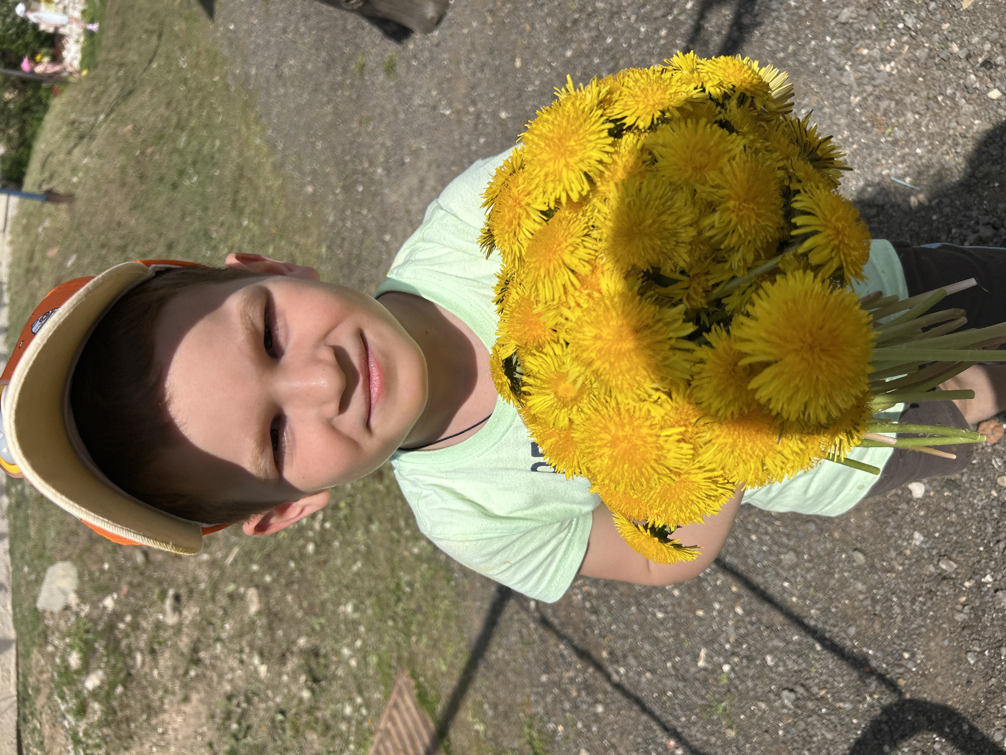 A young boy wearing an orange and white baseball cap and a light green t-shirt is holding a large bouquet of yellow dandelions. The photo is taken from above, looking down at the boy. The background appears to be an outdoor area with grass and pavement. The location is identified as Noviy, Russia. The boy is smiling with his eyes closed, suggesting he is pleased with the bouquet. The time of day appears to be sunny.