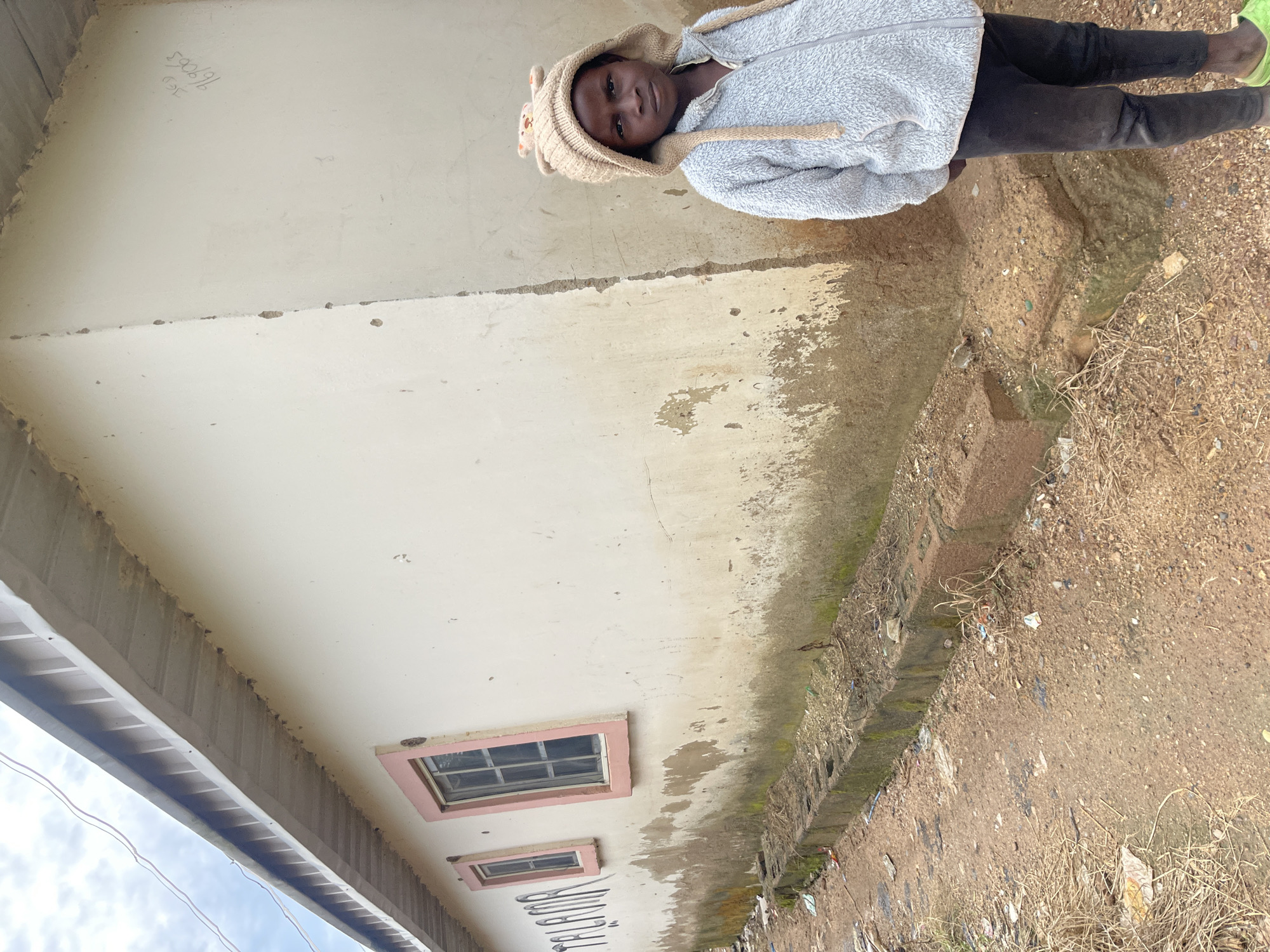 In Jos, Nigeria, a young child stands on a dirt road in front of a partially damaged, light-colored building. The building's exterior wall is stained with mud and grime, and there are some markings on the wall. The roof appears to be made of corrugated metal. Two windows with pink frames are visible on the wall. The child is wearing a light gray hooded jacket with a bear-face design on the hood and black pants. The ground is covered with dirt, scattered debris, and some dry vegetation. A low concrete structure runs along the base of the building. The sky is visible above the building, with some clouds.