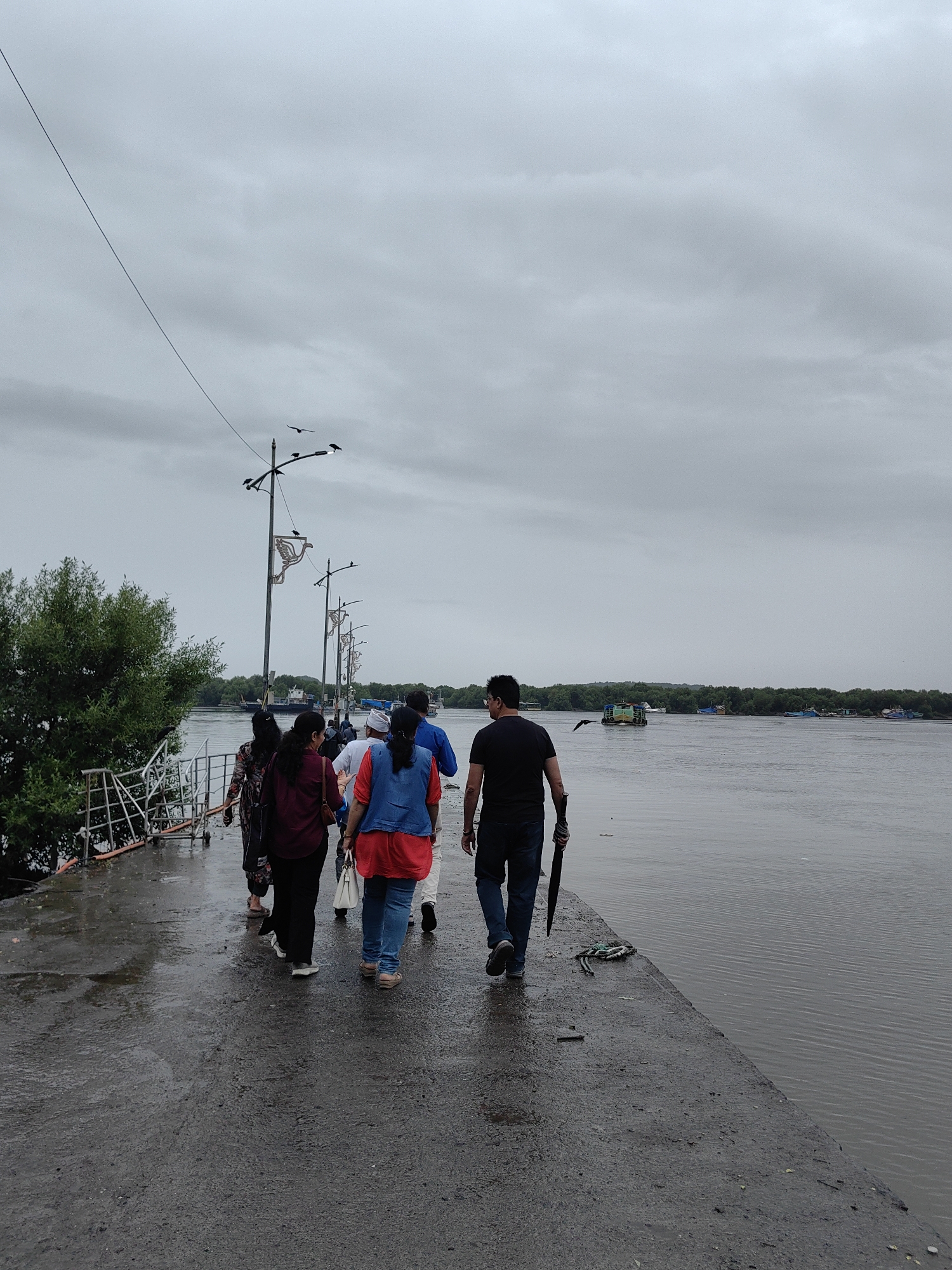 A group of people are walking along a wet concrete pier in Gorai, India. The sky is overcast and the water is calm. The pier is lined with lampposts and a railing. A man in the foreground is carrying a black umbrella. In the background, several boats are visible on the water, and trees line the shore. The overall atmosphere is subdued, suggesting a rainy or humid day.