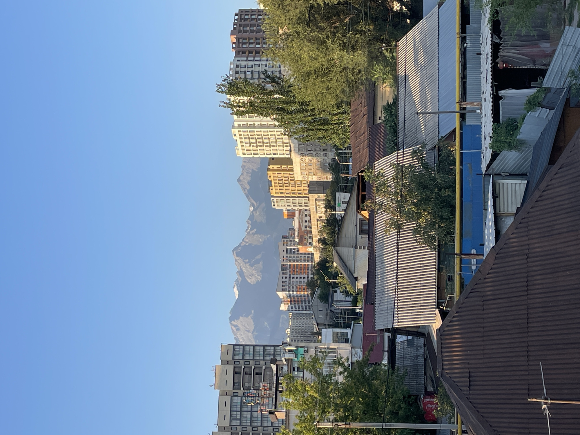 The image is a medium shot, taken from an elevated position, showing a cityscape with mountains in the background. The sky is clear and blue. In the foreground, there are several rooftops of houses, some made of corrugated metal. Behind these houses, there is a line of trees. Further in the background, there are several apartment buildings of various heights and colors, with modern architecture. The tallest buildings are on the left and center of the image, and appear to be residential complexes. In the distance, a range of snow-capped mountains is visible, suggesting the location is Almaty, Kazakhstan, which is known for its proximity to the Tian Shan mountain range. The lighting suggests it is either early morning or late afternoon, with the sun casting shadows and highlighting the textures of the buildings and rooftops. There are no people visible in the image.