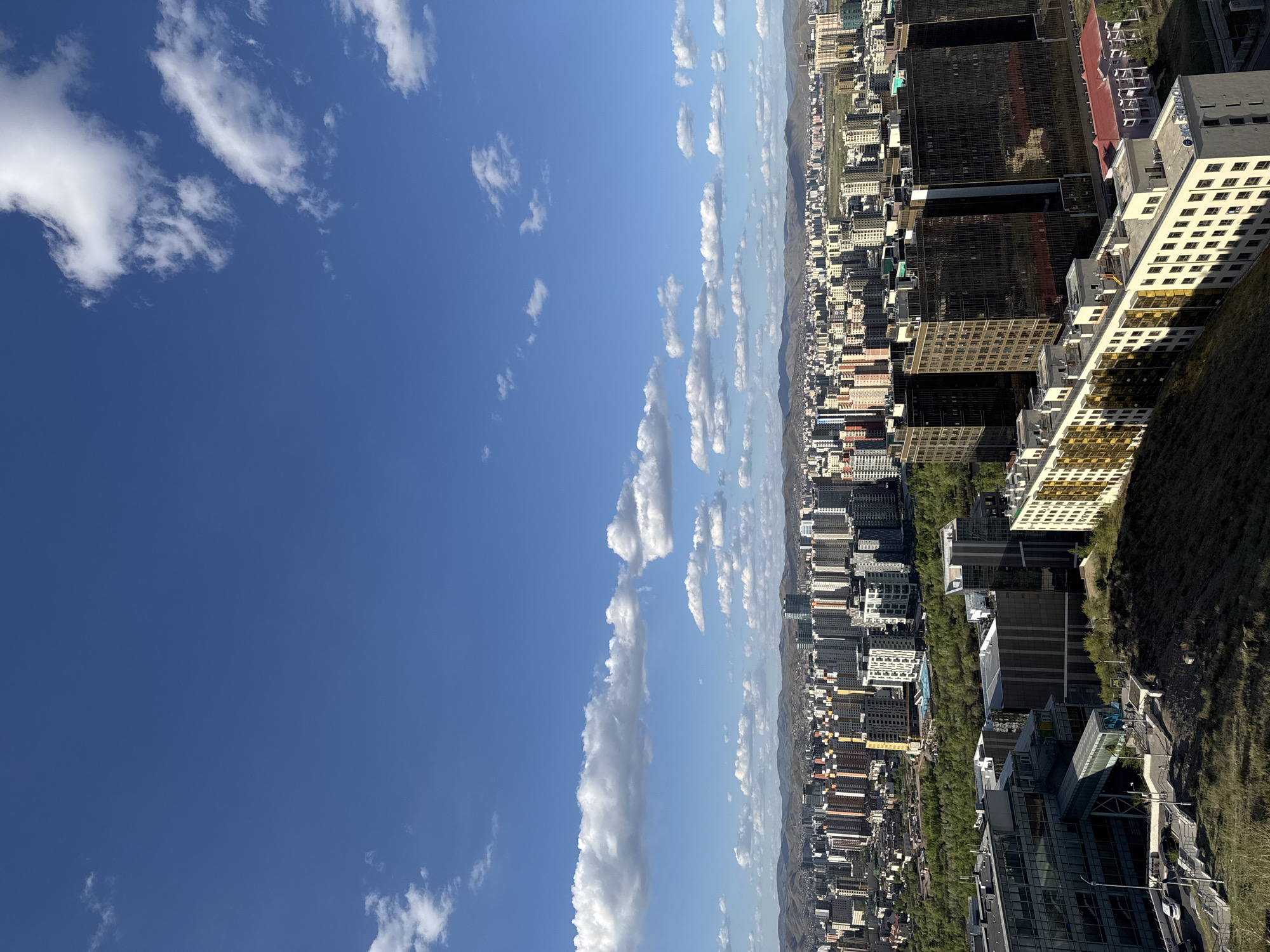 The image shows a partial view of the city of Ulaanbaatar, Mongolia, with a clear blue sky filled with scattered white clouds occupying the left side of the frame. The city extends along the right side, densely populated with various buildings, ranging in height from low-rise to skyscrapers. These structures display diverse architectural styles and colors. Green spaces and trees are visible between buildings, adding to the urban landscape. The horizon line is marked by distant mountains under a slightly hazy sky, creating a visual contrast between the clear sky and the bustling urban environment.