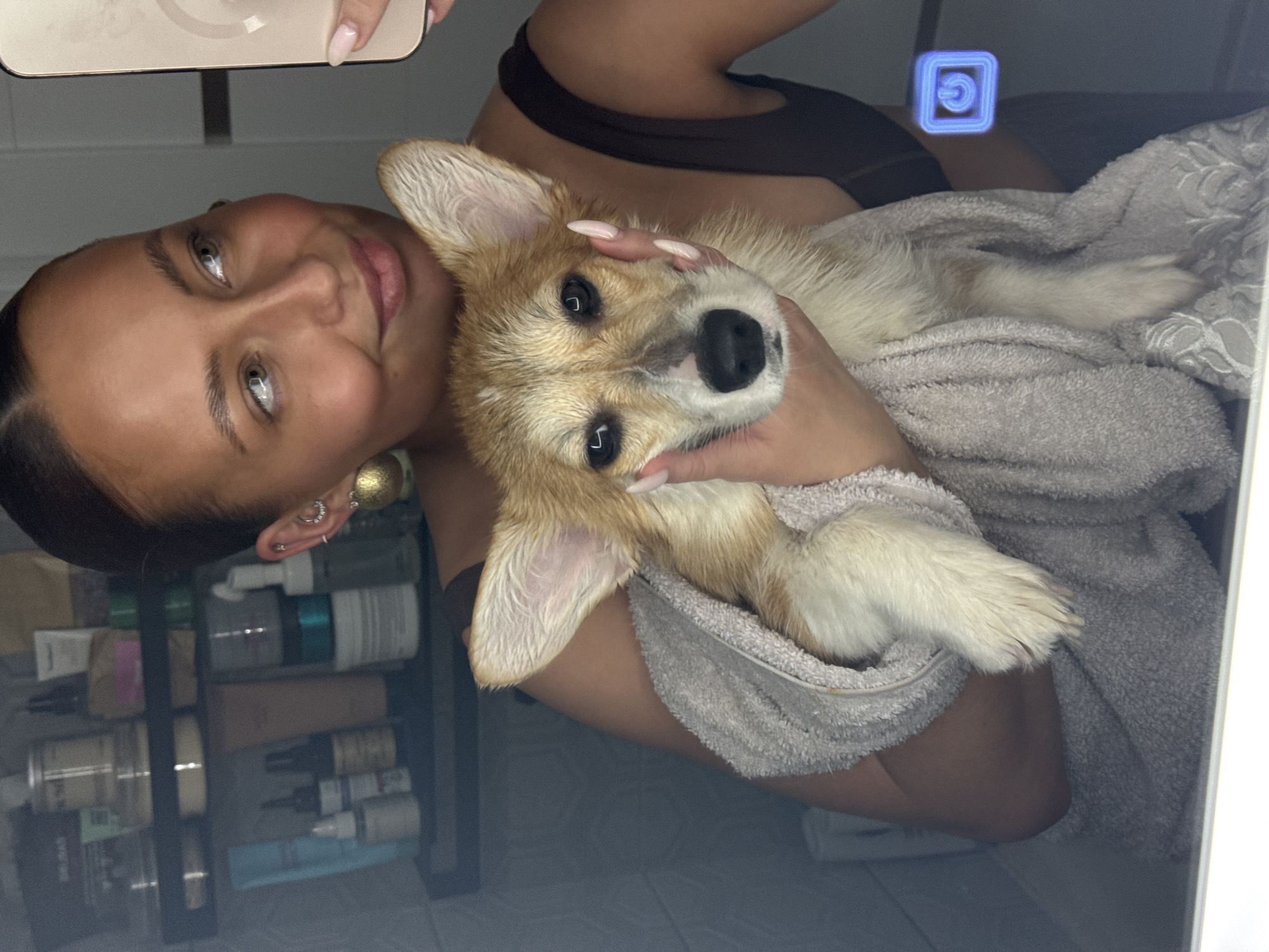 A woman is holding a corgi puppy wrapped in a grey towel, posing for a selfie in a bathroom. The woman has dark hair pulled back, tan skin, and is wearing a dark top. She's holding a phone in her left hand to take the picture. In the background, a shelf unit filled with toiletries is visible. The puppy appears to have wet fur. The location is likely a private residence but no evidence of the city, Chaikovsky, Russia, is present.