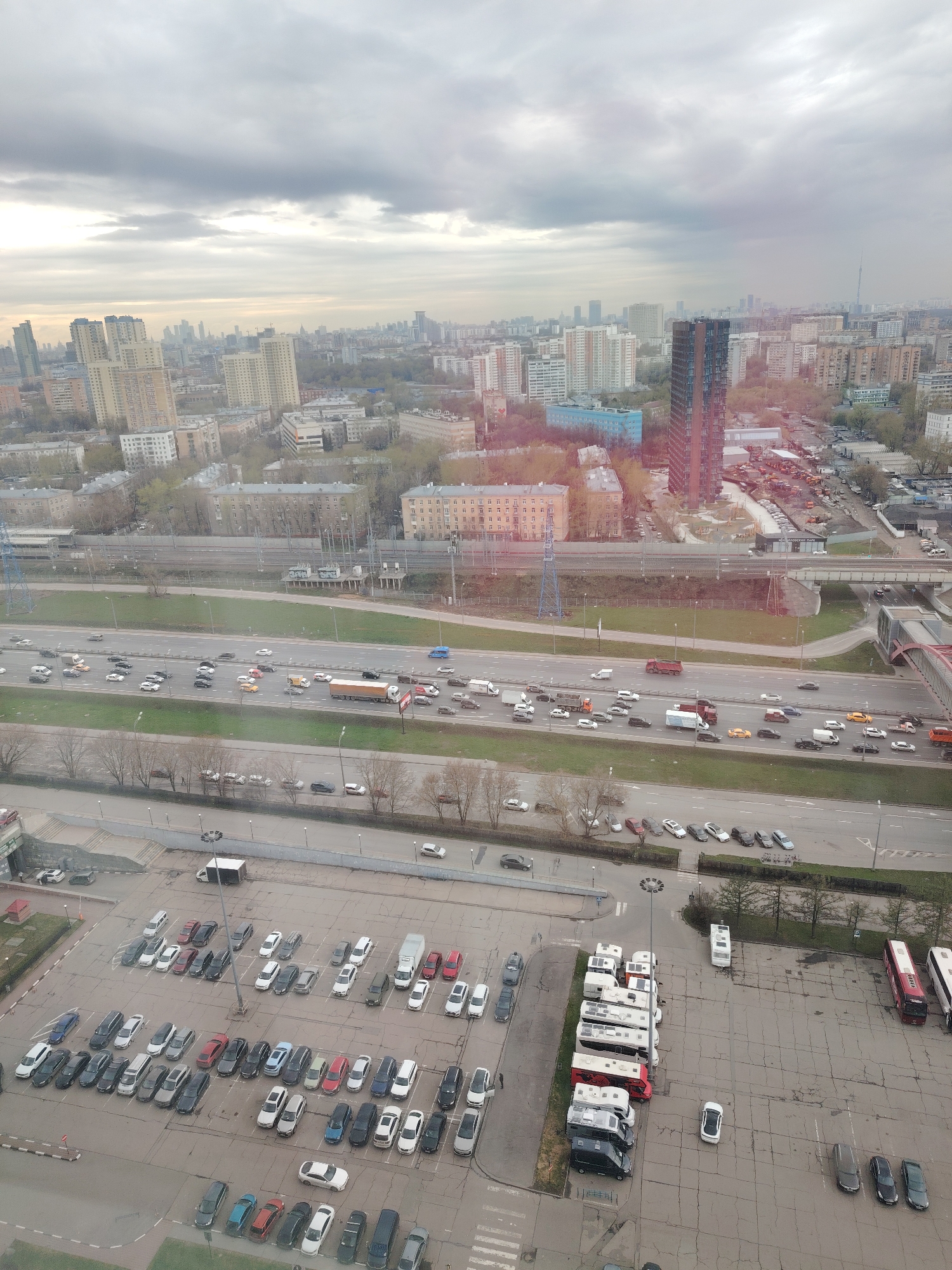 This media file presents a high-angle, expansive view of an urban landscape under an overcast sky.
**Subjects:** The scene is dominated by a city with numerous multi-story buildings, including both older residential blocks and modern high-rises. A wide, multi-lane highway traverses the mid-ground, paralleled by a railway track. Numerous vehicles, including cars, trucks (some semi-trailer trucks, others box trucks), and a concentration of recreational vehicles (RVs) and buses in a large parking lot, are visible. Bare trees line the roads and parking areas. In the far distance, a very tall, slender structure, possibly a communication tower (resembling the Ostankino Tower), is faintly visible on the horizon. A distinctive tall, dark, and slender skyscraper stands prominently in the mid-right.
**Activity/Event:** The primary activity is vehicle movement on the highway, indicating daily traffic. Vehicles are also parked in various lots, suggesting routine urban parking.
**Notable details:** The time of day appears to be daytime, likely mid-day, given the diffuse light and absence of long shadows. The weather is heavily overcast, with a sky full of uniform grey clouds. The architectural styles, road infrastructure, and the distant tower strongly suggest a large Eastern European city, specifically resembling Moscow. Faint Cyrillic text, possibly "МЕБЕЛЬНЫЙ ПАРК" (Mebelnyy Park - Furniture Park) or similar, is barely legible on a sign near a construction area adjacent to the dark skyscraper. No specific emotions are conveyed by the scene; it depicts a typical, busy urban setting.