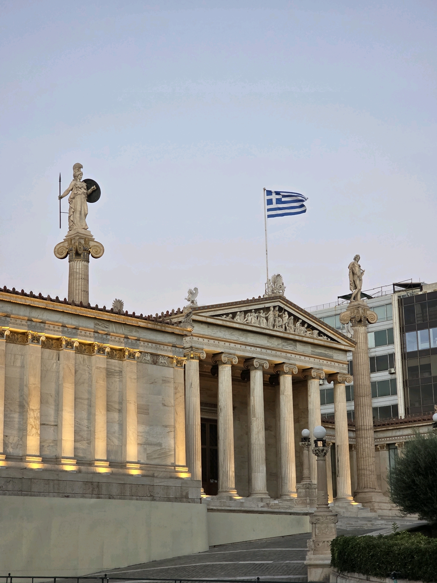 The image captures the exterior of the Academy of Athens in Greece, a neoclassical building with a prominent Greek flag flying. The building is characterized by its white marble facade, ornate columns, and detailed sculptures adorning the pediments. Two statues are visible: one of Athena on a tall column on the left, and another of Apollo on a similar column to the right. The Academy is flanked by modern buildings, contrasting with its classical architecture. The scene is illuminated by warm artificial lights that highlight the marble columns and architectural details, suggesting it is either dusk or early evening. The sky is clear and pale blue.