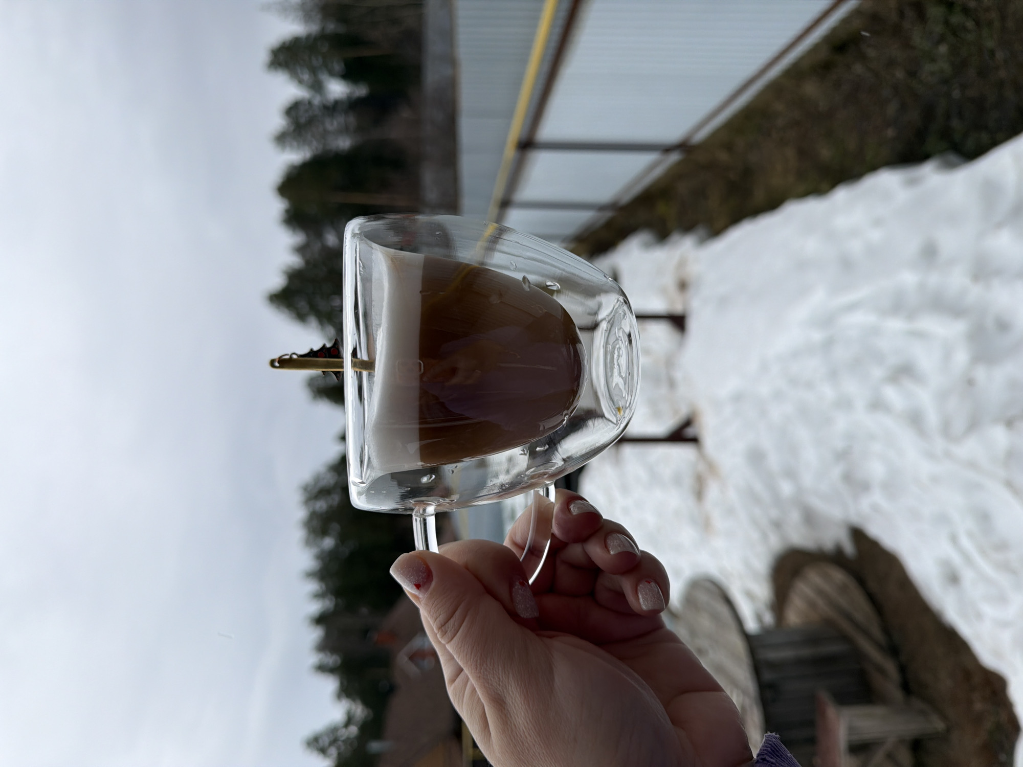 A hand holds a glass of coffee outside in a winter setting. The glass, with a transparent double-wall design, contains a brown liquid that appears to be coffee. The hand holding the glass has nails with a subtle glittery polish. The background shows a scene covered in snow, with trees visible in the distance. A wire fence is also visible, suggesting the location is possibly near a residential area or property boundary. The presence of snow and the cloudy sky suggests a cold, possibly overcast day. It is not possible to confirm the location is the city of Chaikovsky, Russia from the image itself.