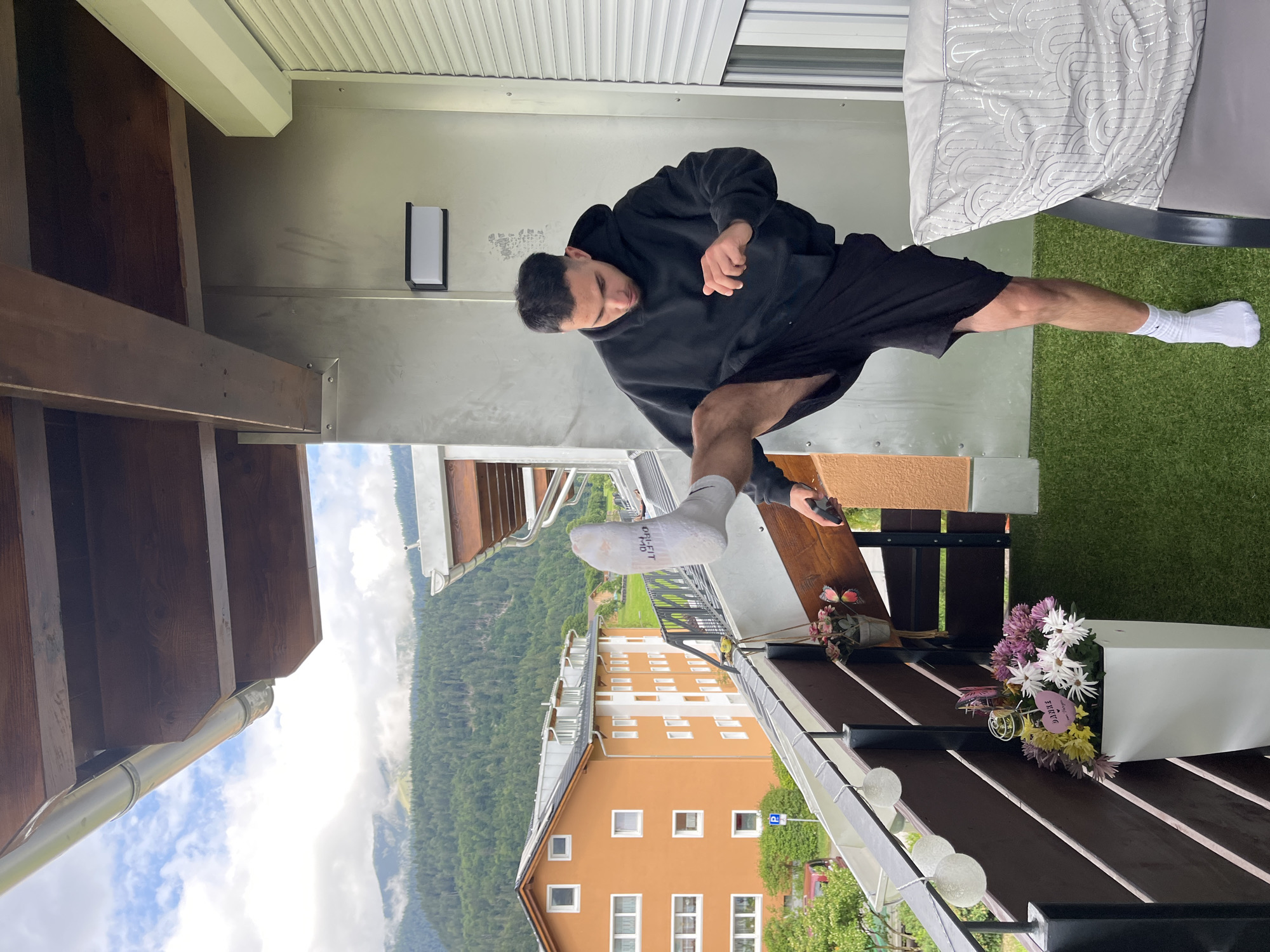 The image depicts a male individual performing a stretch on a residential balcony in Garmisch-Partenkirchen, Germany. The man, with short dark hair and stubble, is dressed in a black oversized hoodie, black shorts, and white socks. His left leg is raised high, with the sole of his left sock visible, displaying "DRI-FIT MD" text. His left hand holds a dark object, possibly a smartphone, near his body. He is positioned on a balcony featuring artificial green turf flooring.
The balcony railing is composed of dark metal posts and horizontal wooden planks, with a string of spherical white lights draped along the top. A tall, white rectangular planter containing a variety of white, purple, and yellow flowers is situated on the floor near the railing. A pink heart-shaped tag, inscribed with "DANKE," is embedded among the flowers. A small pot with succulents hangs from a wooden beam above the railing. The balcony ceiling consists of dark wooden beams and planks, beneath which a light-colored rain gutter is visible. A modern rectangular black wall light with a white diffuser is mounted on a light grey wall to the right of the man. Further right, a light-colored roller shutter (Jalousie) covers a window or door opening. A grey outdoor chair with a patterned cushion (silver arcs on grey/white) is partially visible on the far right.
Beyond the balcony, a multi-story building with an orange facade and numerous white-framed windows is prominent. Another building with a white roof and rain gutters is visible to the right of the orange building, further in the background. Green foliage, including trees and bushes, is present around the buildings and along a black metal fence. A blue parking sign ("P") is discernible near a road below. In the distant background, densely forested mountains rise under a partially cloudy sky.