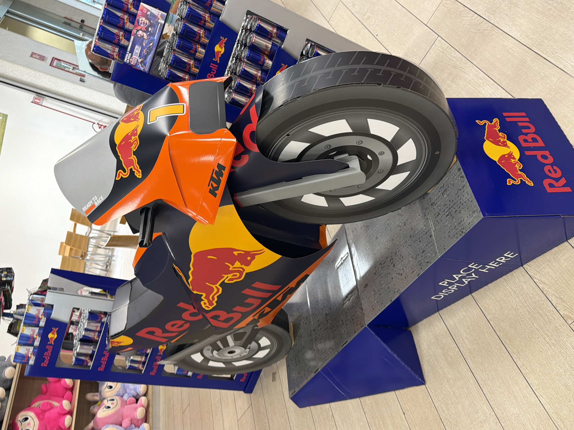 In the city of Due Carrare, Italy, a Red Bull branded motorcycle cutout is displayed on a blue pedestal. The cutout is orange and black, featuring the Red Bull logo and the number "1". Behind the motorcycle, shelves are stocked with Red Bull cans. The environment appears to be a retail store, with wooden flooring visible. No people are present in the immediate foreground of the image.
