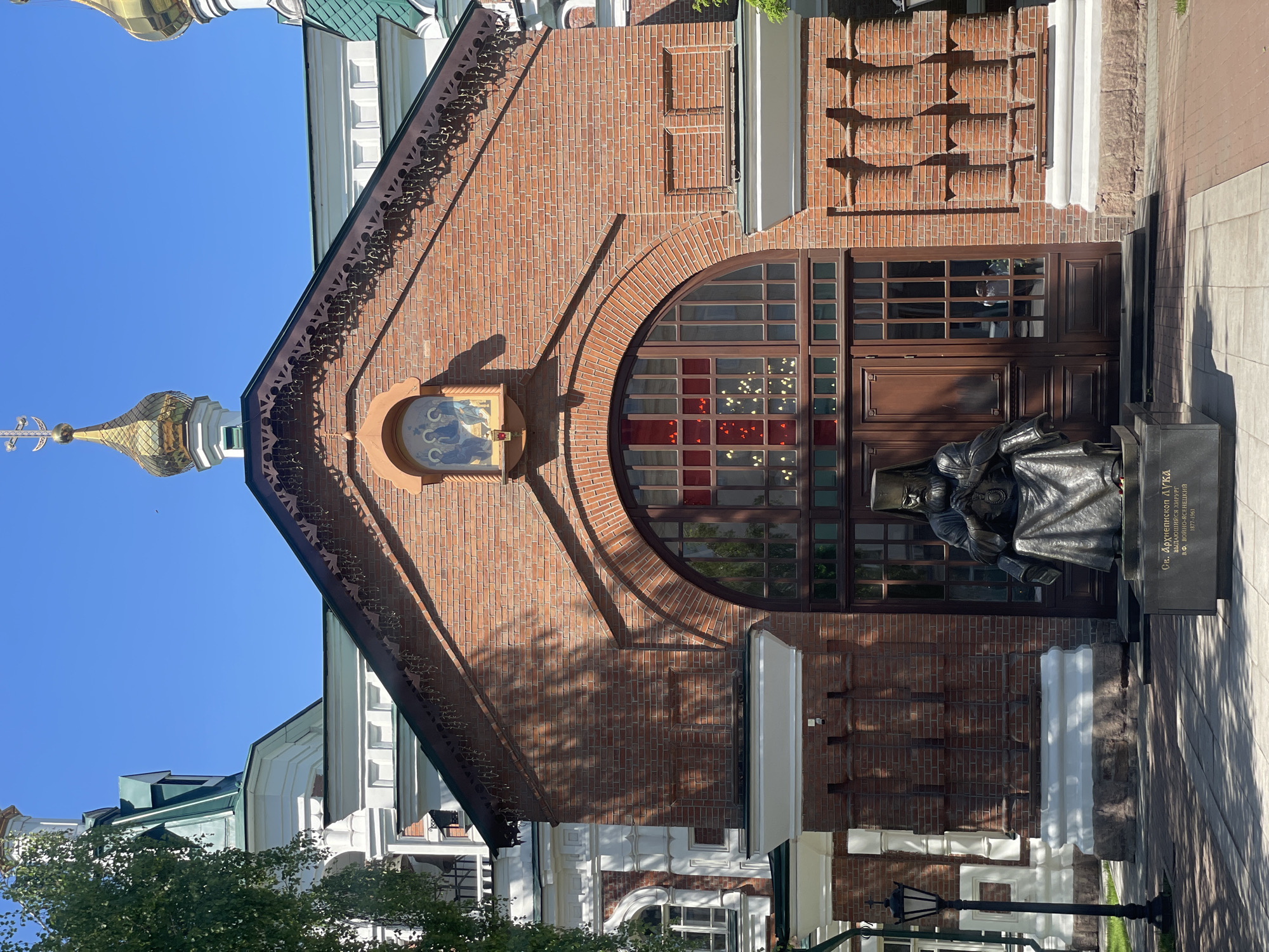 An image taken on a bright, sunny day showcases the exterior of a red brick building, likely a church or religious institution, with clear blue sky above. To the left, a golden onion dome topped with a cross is visible. The building features an ornate dark green roofline with decorative trim.
The main facade is constructed from red bricks, with a large, arched window dominating the center. This window has wooden frames and contains stained-glass-like panels, some clear and others with red sections. Above this arched window, set into the brickwork, is a religious icon depicting figures, possibly the Virgin Mary and Child. To the right of the window is a wooden double door with intricate paneling.
In front of the building, on the right side, stands a dark-colored statue of a robed figure, seemingly seated or kneeling, looking down at a book placed on a black pedestal. The pedestal bears Cyrillic text which reads: "Св. Архиепископ ЛУКА" (St. Archbishop LUKE), and below that "Выдающийся хирург" (Outstanding surgeon), "И. ВОИНО-ЯСЕНЕЦКИЙ" (I. Voino-Yasenetsky), and the dates "1877-1961". This identifies the statue as Saint Luke of Simferopol, who was also a surgeon. Strong shadows from the sun indicate a bright day, possibly morning or afternoon. A traditional street lamp is partially visible on the far left. The overall setting appears to be urban, in Krasnoyarsk, Russia.
