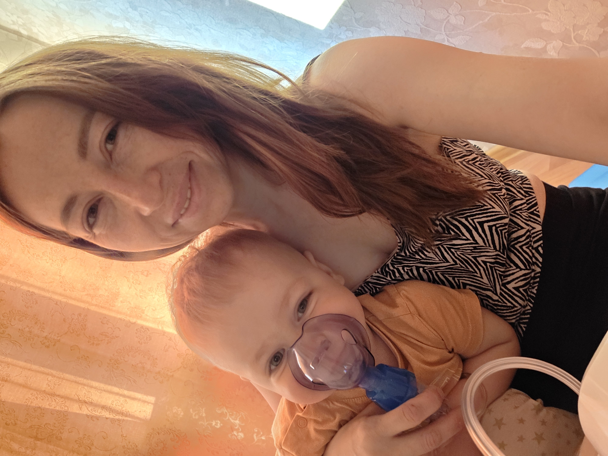 A woman is holding a young child with a nebulizer mask on. The woman is smiling and appears to be taking a selfie. The child is looking at the camera. The environment has patterned wallpaper. The location is unspecified, but the city is Chaikovsky, Russia.