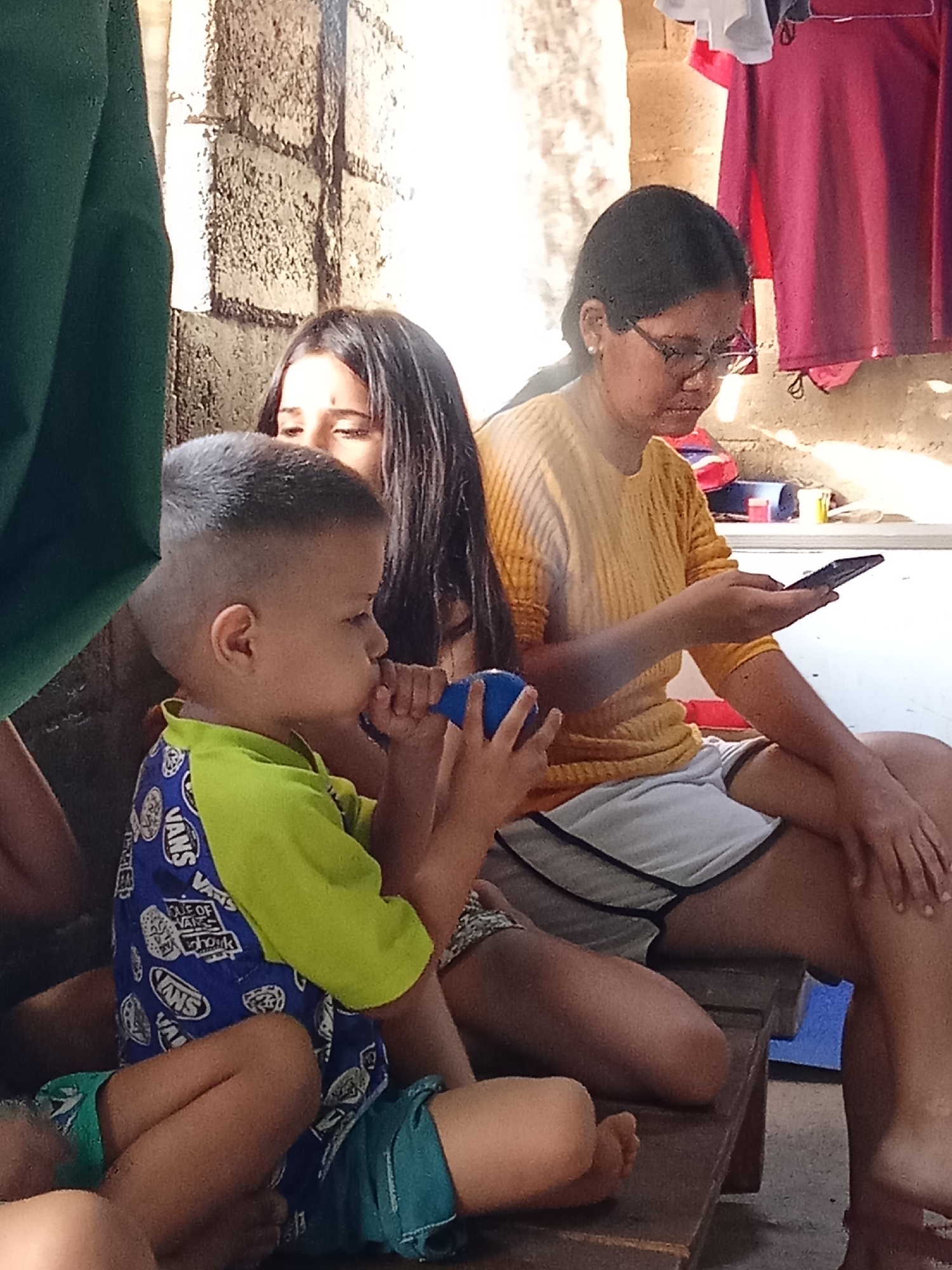 Three people are sitting on a wooden bench outdoors. A young child is blowing into a blue toy instrument. An older girl with long black hair is sitting next to the child. An adult woman wearing glasses and a yellow shirt is looking at a cell phone. A concrete wall is in the background. A piece of clothing is hanging behind the woman. Based on the information provided, the location is Juan Griego, Venezuela.