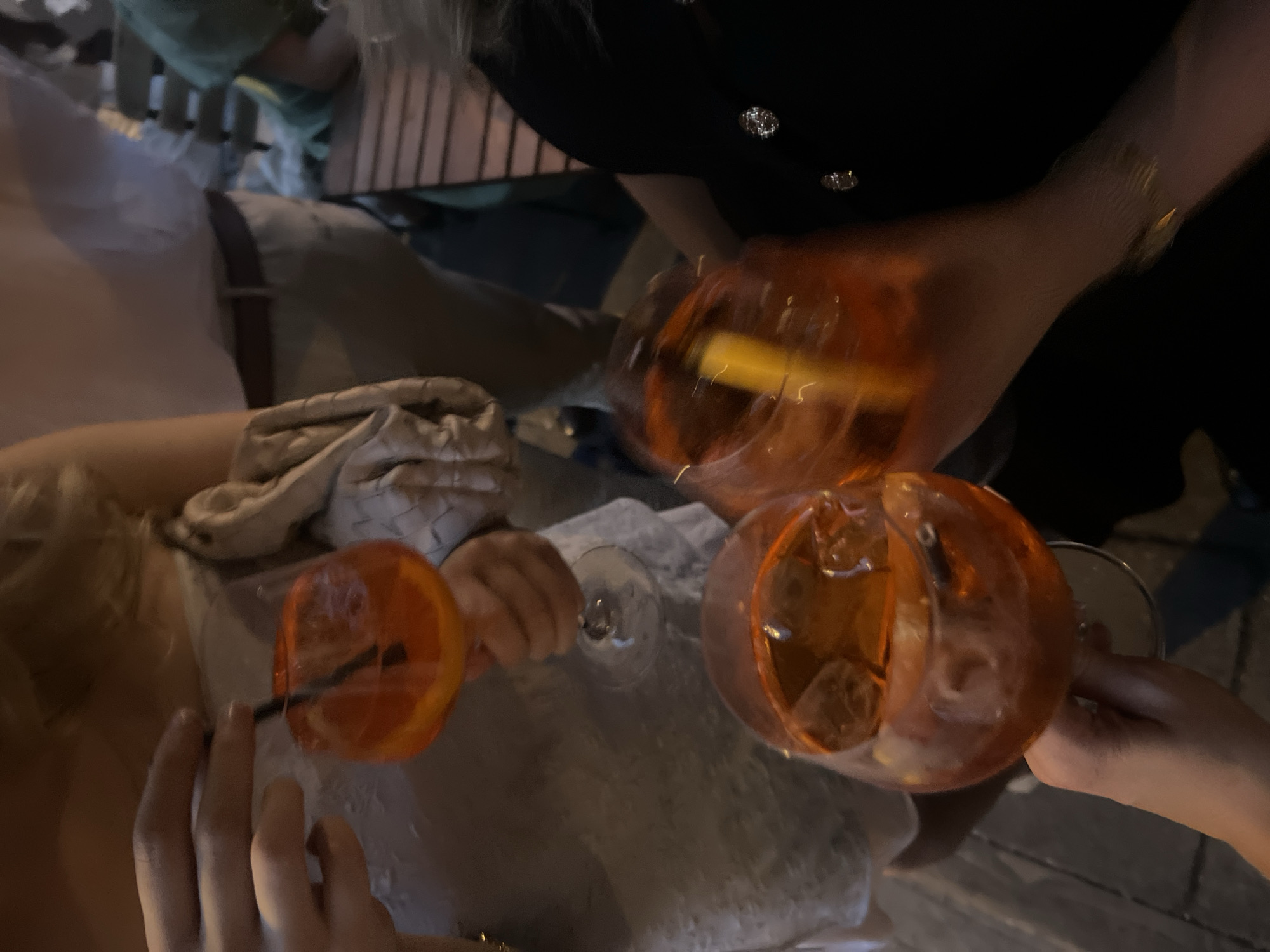 The image depicts three individuals toasting with orange-colored beverages in round glasses, likely Aperol Spritz cocktails, with ice cubes and garnishes of orange slices. Two of the individuals are holding glasses, one with a straw, while the third individual is slightly blurred, but also holding a glass. The location appears to be a restaurant or bar, suggested by the presence of tables and chairs. The ambient lighting is dim, indicating an evening or nighttime setting. No specific landmarks or indicators confirm this is London, United Kingdom.