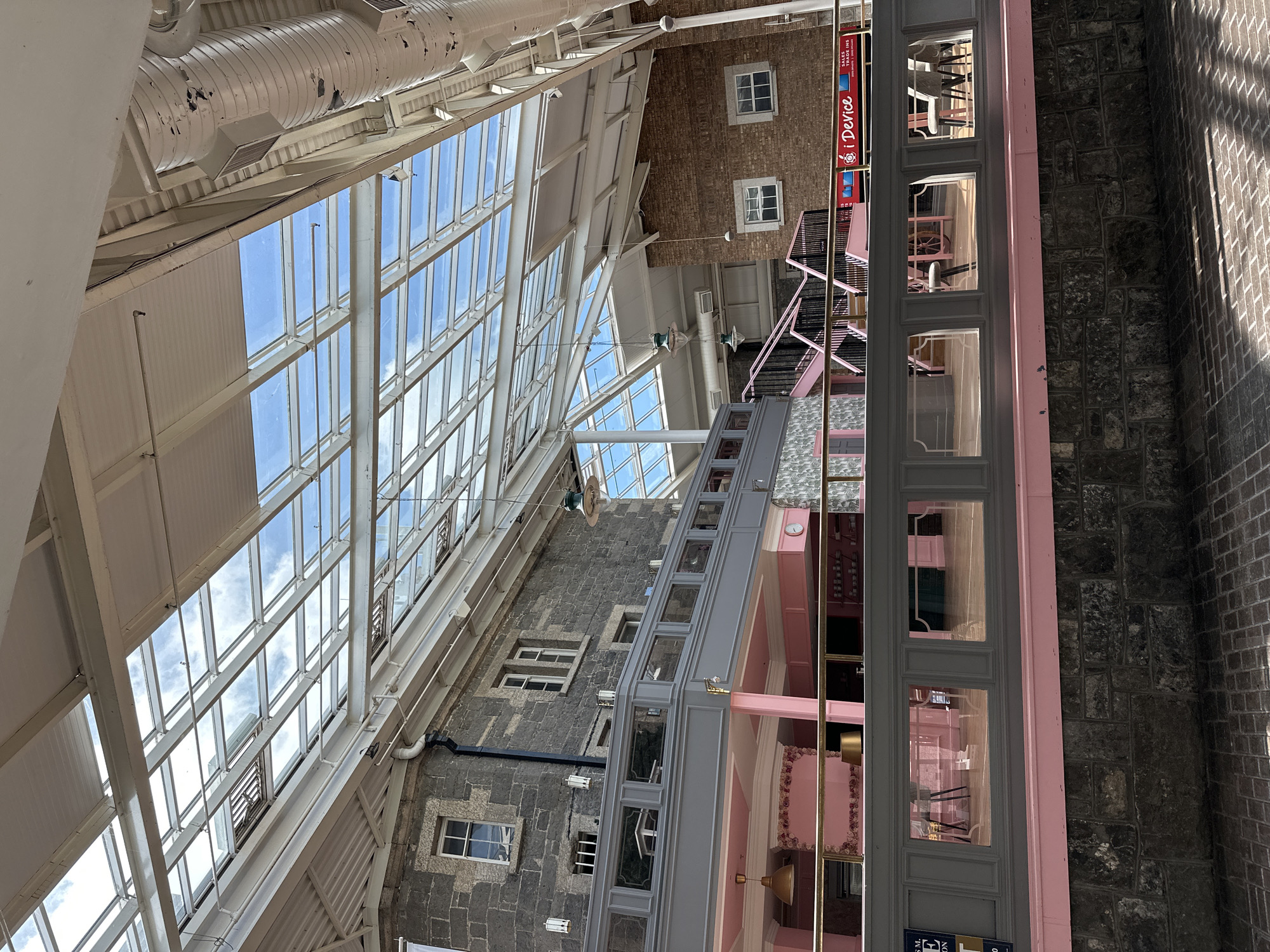 The image depicts the interior of an atrium with a large glass roof. Sunlight streams through the roof, illuminating the space and casting shadows on the brick floor. On the left side of the image, a grey and pink balcony overlooks a pink-themed interior with tables and chairs. A stone building with windows is visible behind the balcony. On the right side, a brick building with windows and a red "i Device" sign is present. A set of pink stairs with black railings leads up to a higher level. The overall scene suggests an indoor market or shopping center. The location is identified as Carlow, Ireland, though no specific landmarks are visible to confirm this. There are no people visible in the image.