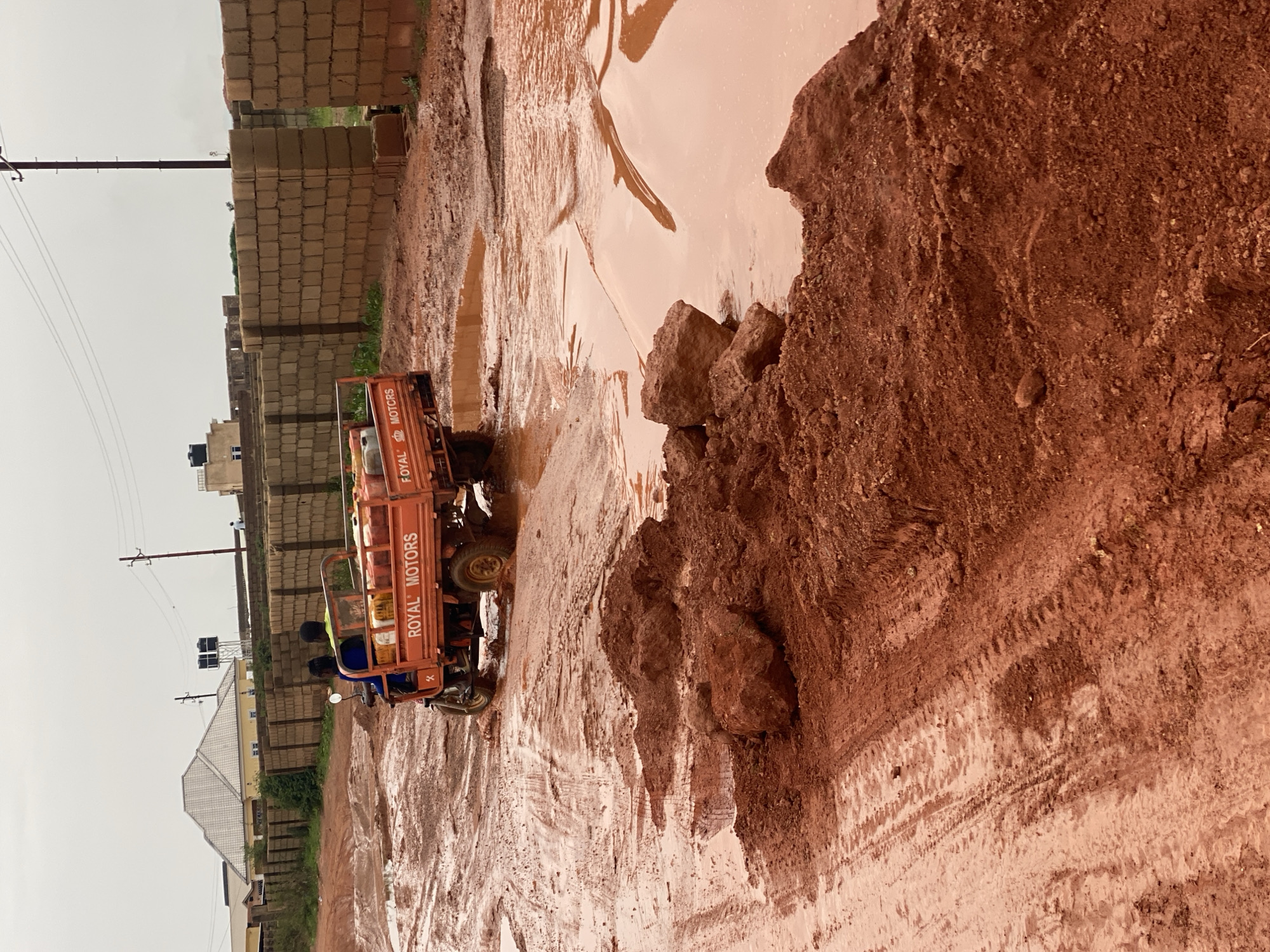The image depicts a scene in Jos, Nigeria, likely after heavy rainfall. A red, three-wheeled vehicle, possibly a utility tricycle, is partially submerged in a muddy puddle on an unpaved road. The vehicle has "ROYAL MOTORS" printed on its side. Two individuals are visible on the vehicle, appearing to navigate the challenging terrain.
The surrounding environment is characterized by large piles of reddish-brown soil and bricks, suggesting a construction site or a quarry. The ground is heavily saturated with water, forming large, murky puddles that reflect the overcast sky. The weather appears to be gloomy, indicated by the muted light and the general atmosphere of the scene.
In the background, a line of brick walls and some structures, including a house with a gabled roof and what appear to be water tanks on a building, are visible. Telephone poles with wires crisscross the sky, indicating the presence of infrastructure. The overall impression is one of a difficult and potentially hazardous environment, likely a rural or semi-urban area affected by poor road conditions and weather. The activity captured is the movement of the vehicle through the flooded road, highlighting the impact of the elements on daily life and transport.