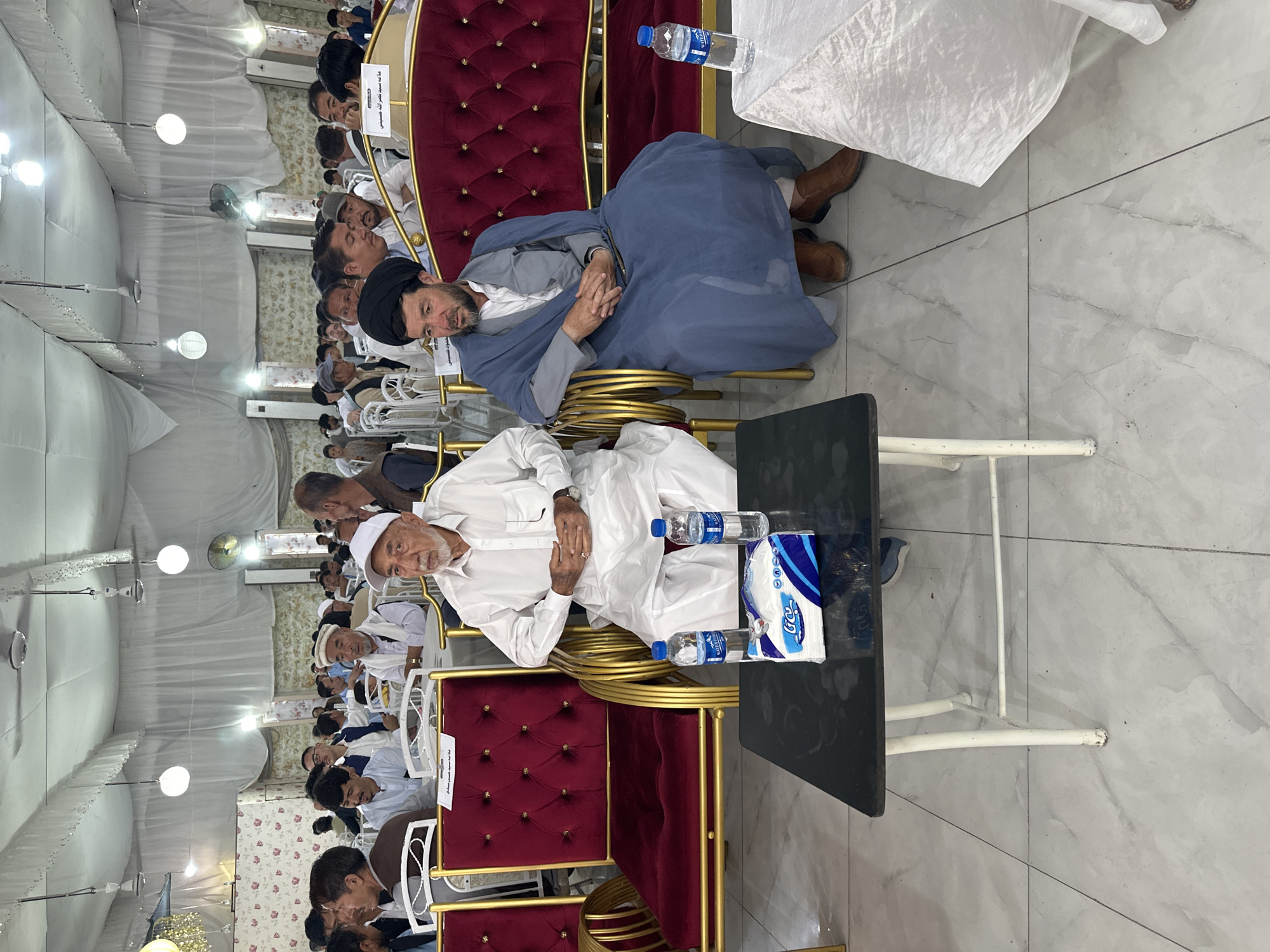 The image depicts a gathering of people inside a tent-like structure in an unknown city, Unknown. Many attendees are seated on ornate red and gold chairs. An elderly man in white clothing is positioned towards the front, near a black table containing water bottles and a package of tissues. Nearby, another man wearing a black turban and a blue garment reclines. Other individuals are seated behind them, with numerous lights and fans visible along the tent's roof.